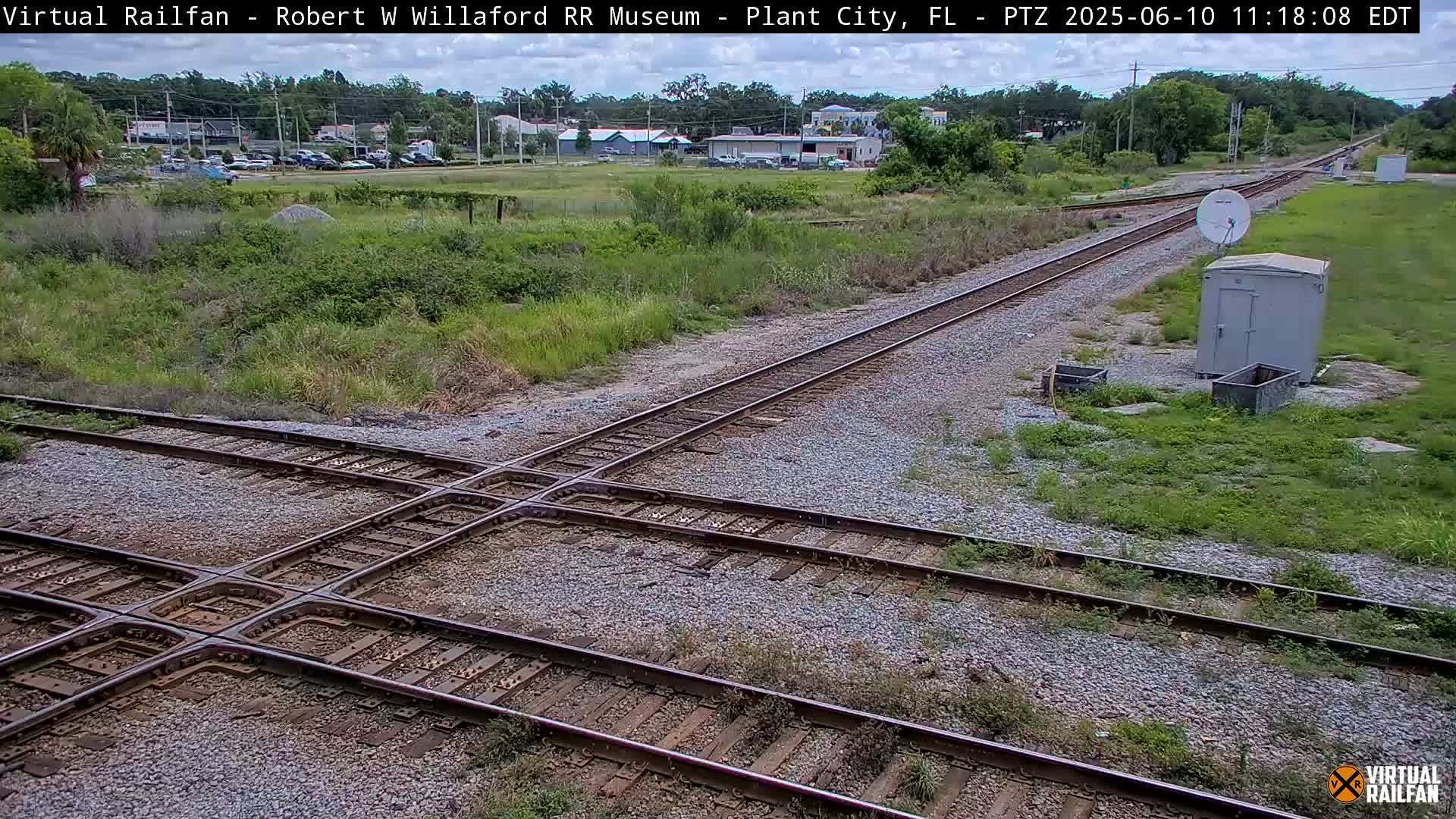 A railroad crossing with two sets of tracks intersecting, surrounded by patchy grass and a small, gray shed under a partly cloudy sky.