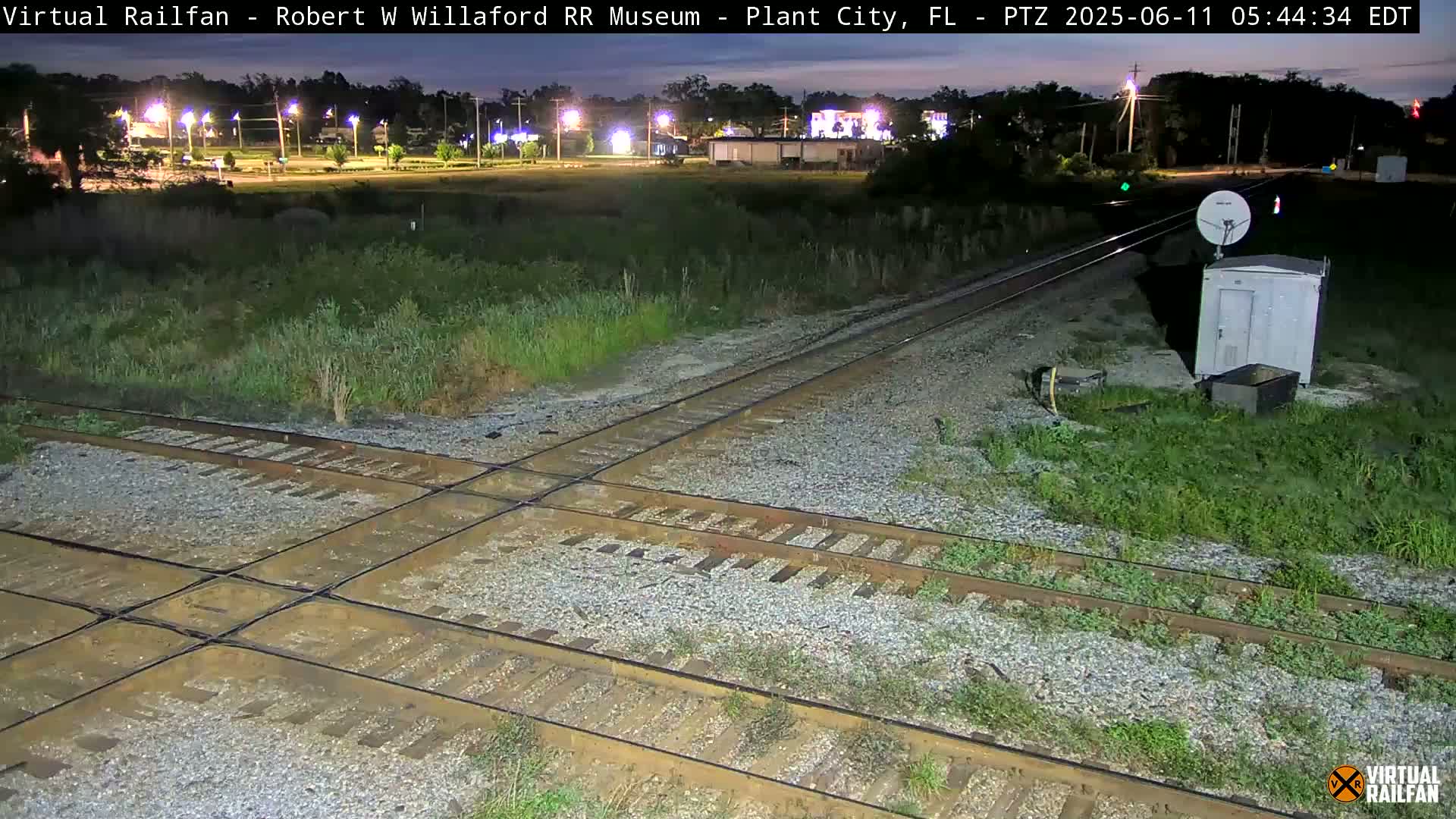 A nighttime view of a railroad crossing with intersecting tracks, surrounded by overgrown vegetation and a small building with a satellite dish.