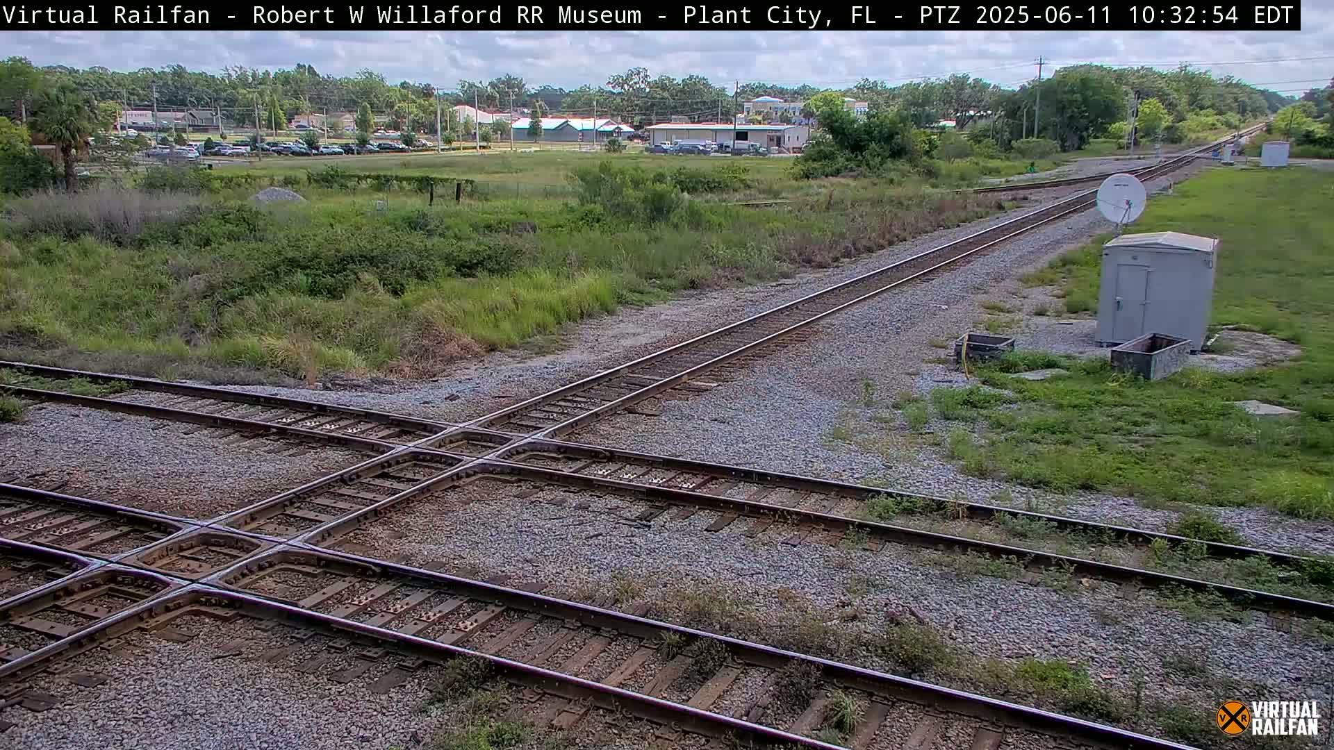 A railroad crossing with several tracks intersecting, surrounded by grassy areas and a small building, under a partly sunny sky.