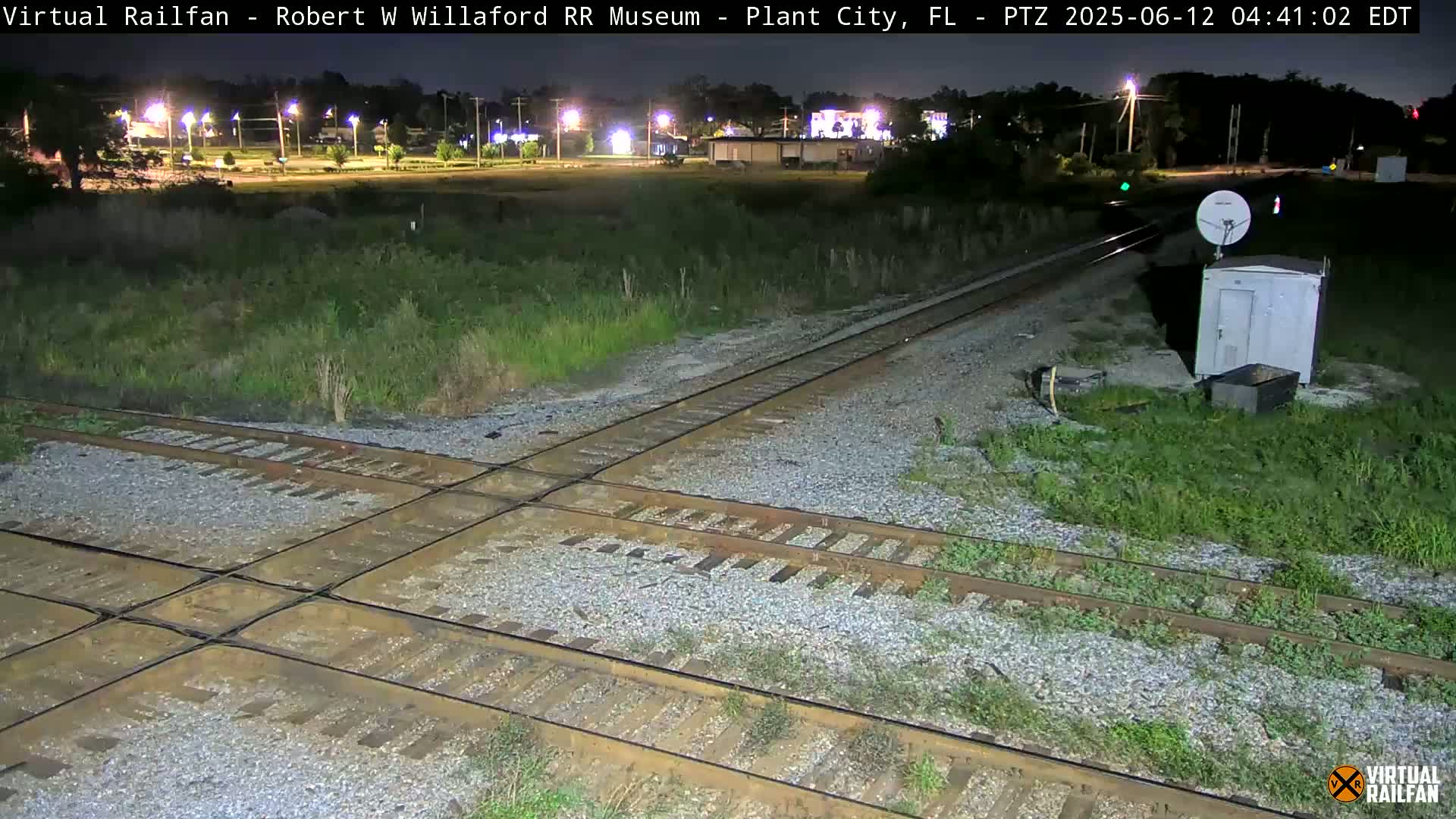 A nighttime view of a railroad crossing with intersecting tracks, surrounded by overgrown vegetation and a small building with a satellite dish.