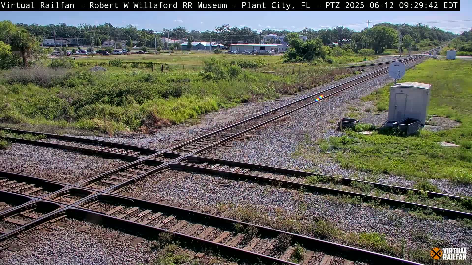 A high-angle, daytime view shows a railroad crossing with two sets of tracks intersecting, surrounded by grassy areas and a small building, under sunny skies.