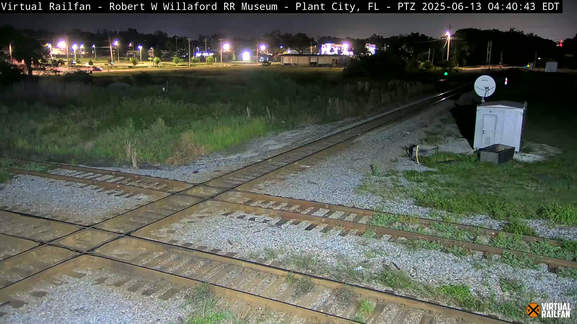 A nighttime view of a railroad crossing with a small building and satellite dish nearby, under clear skies.