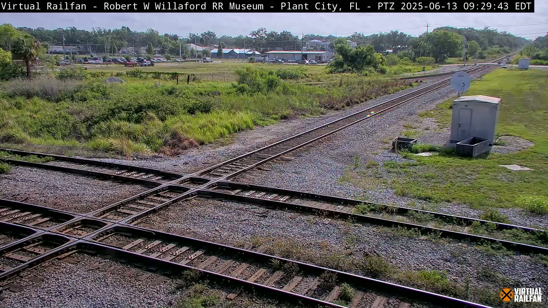 A high-angle, daytime view of a railway crossing with two sets of tracks intersecting, surrounded by grassy fields under a clear sky.