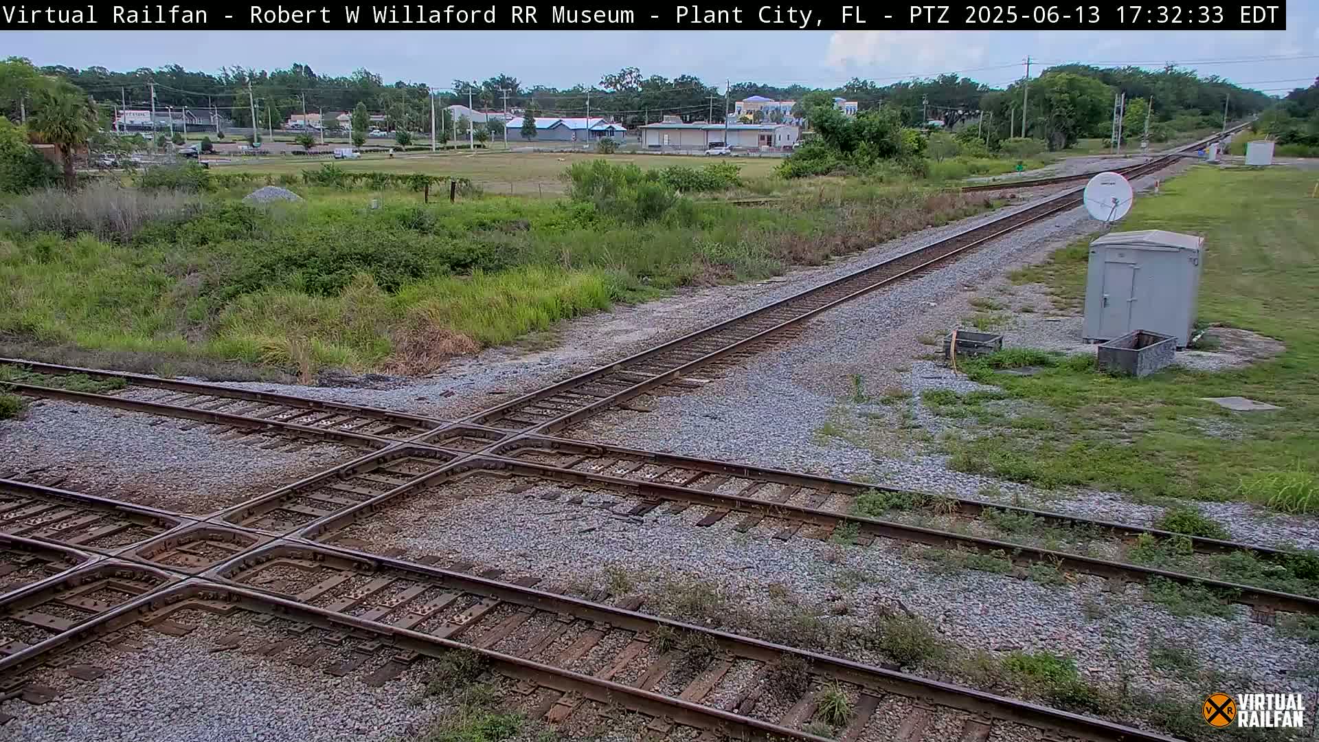 Train tracks intersect at a crossing, with a small building and satellite dish visible in the distance under a partly cloudy sky.