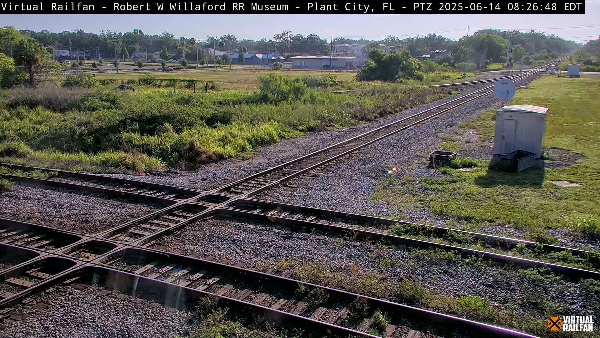 A high-angle view of a railroad crossing on a sunny day, with overgrown vegetation surrounding the tracks.