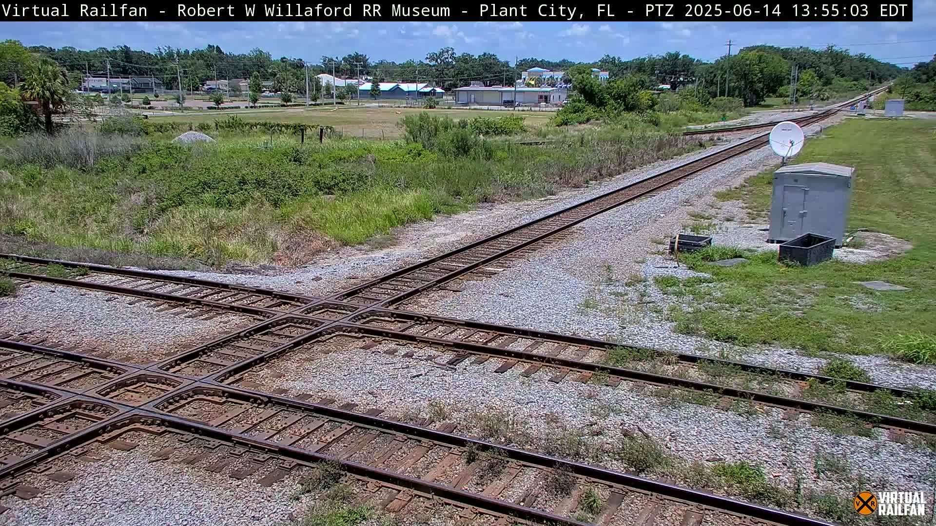 A high-angle, sunny-day view of a railway crossing with two sets of tracks intersecting, surrounded by sparse vegetation and a small utility shed.