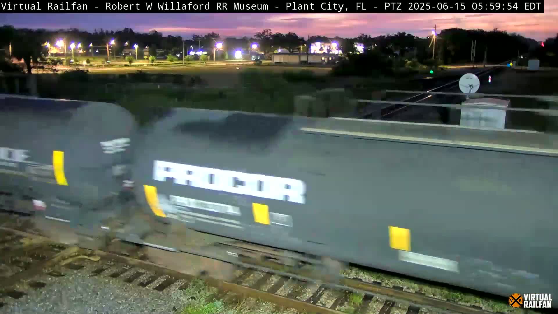 A long freight train passes by at twilight, with the sky showing shades of orange and purple.