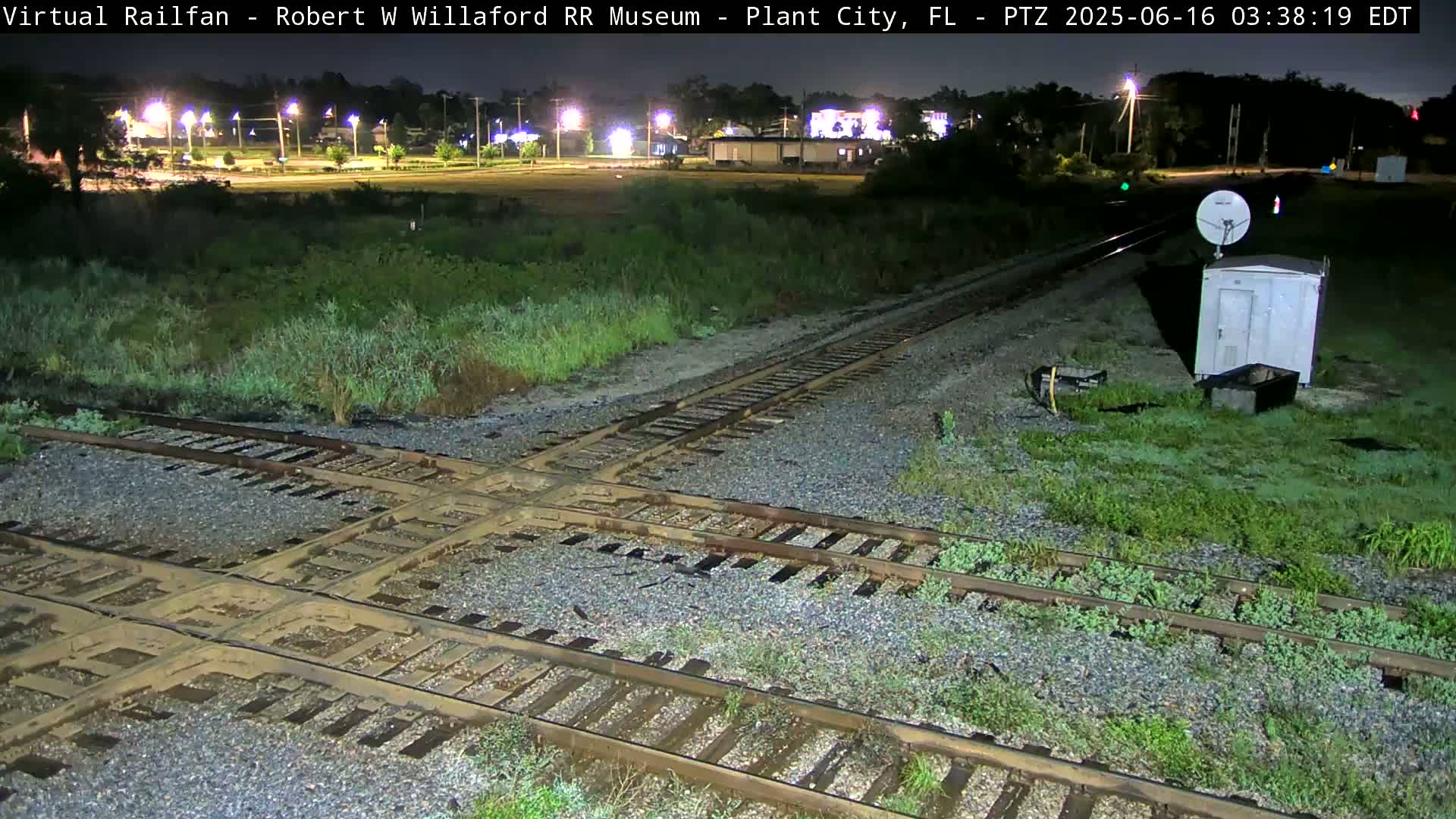 A nighttime view of a railroad crossing with intersecting tracks, surrounded by overgrown vegetation, near a small building and satellite dish.