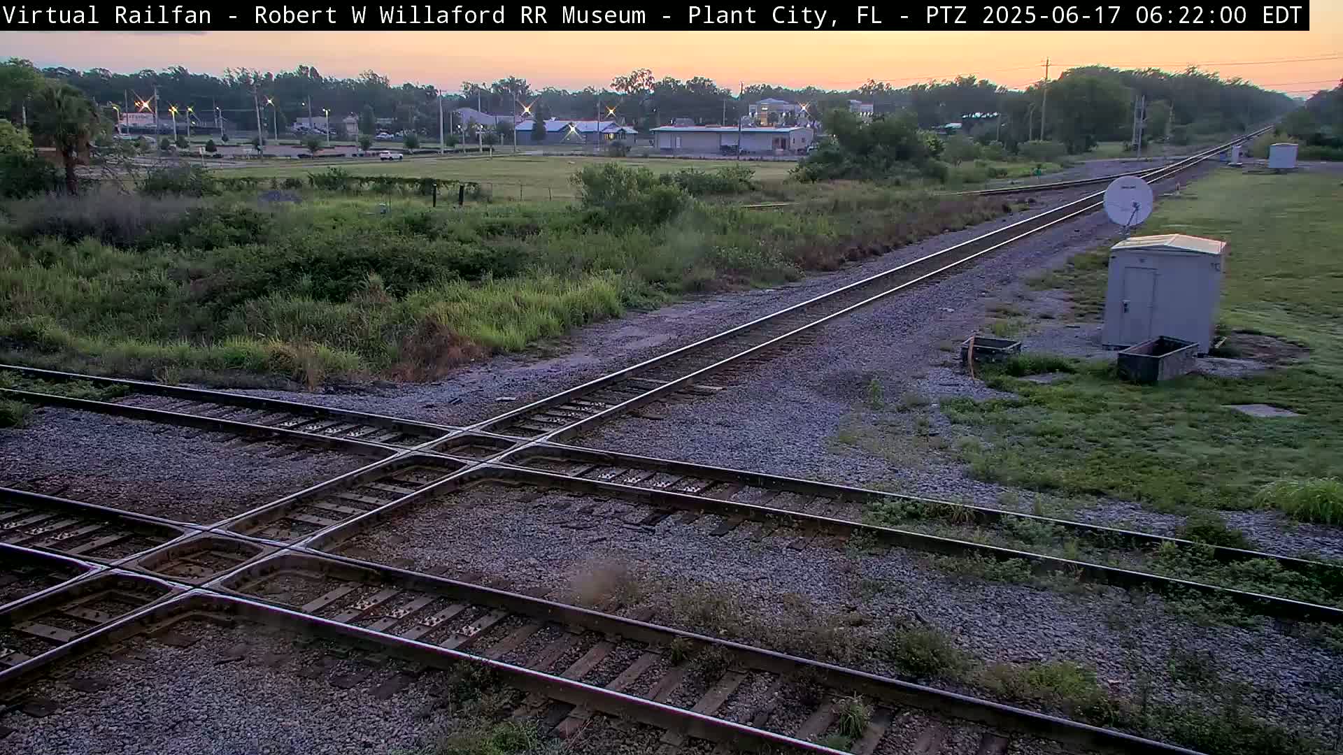 A high-angle view of a railroad crossing at dawn, with two sets of tracks intersecting amidst sparse vegetation and small structures.