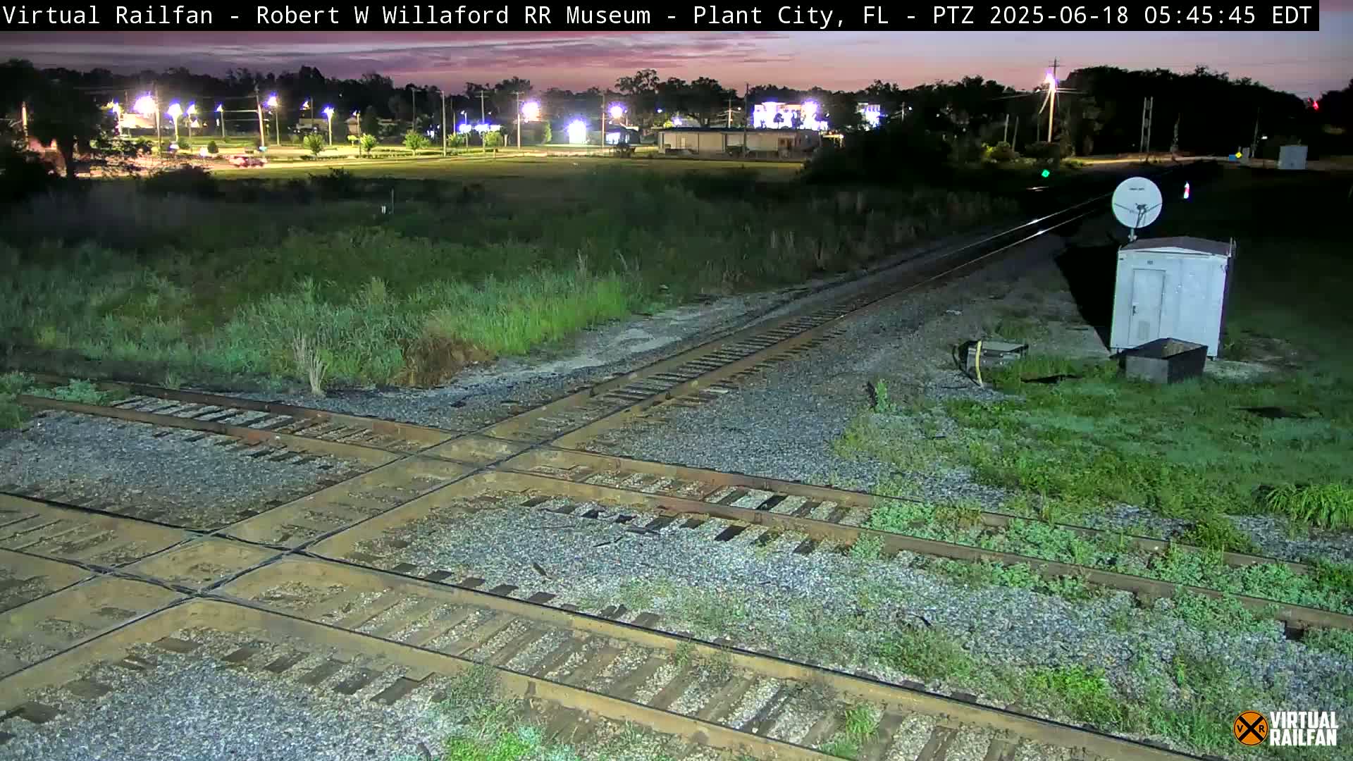 A nighttime view of a railroad crossing with a small building and satellite dish nearby, under a mostly clear sky.