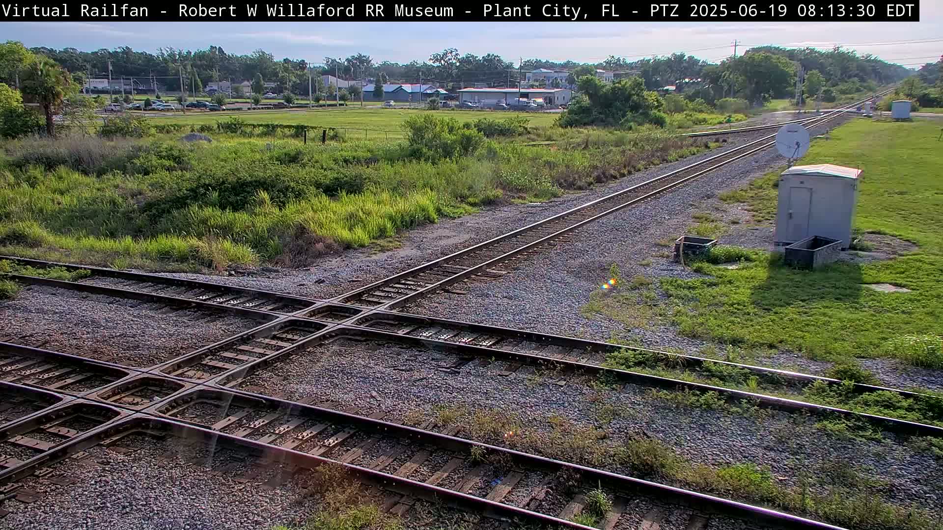 A railroad crossing with intersecting tracks is surrounded by grassy areas and a small building under a sunny sky.