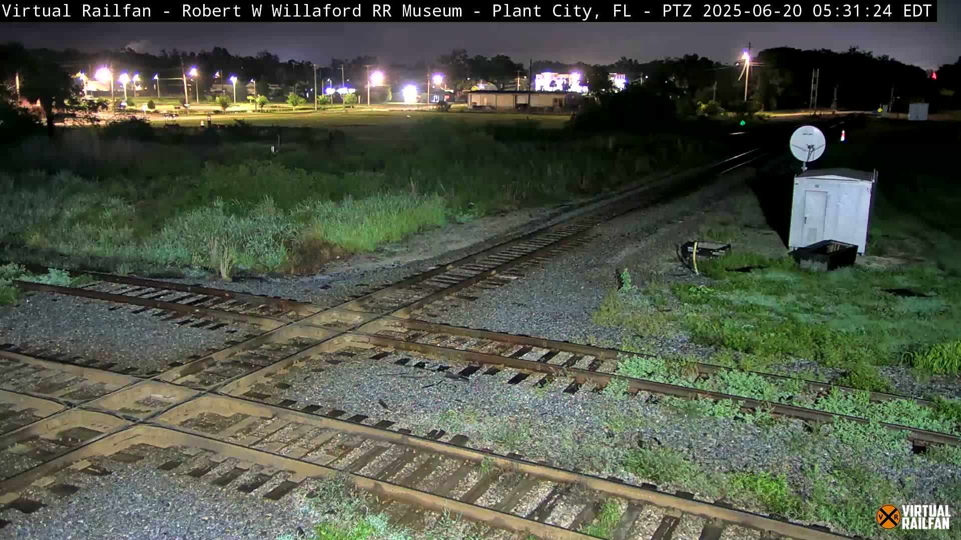 A nighttime view of a railroad crossing with overgrown vegetation, a small building, and a satellite dish, under dark, possibly overcast skies.