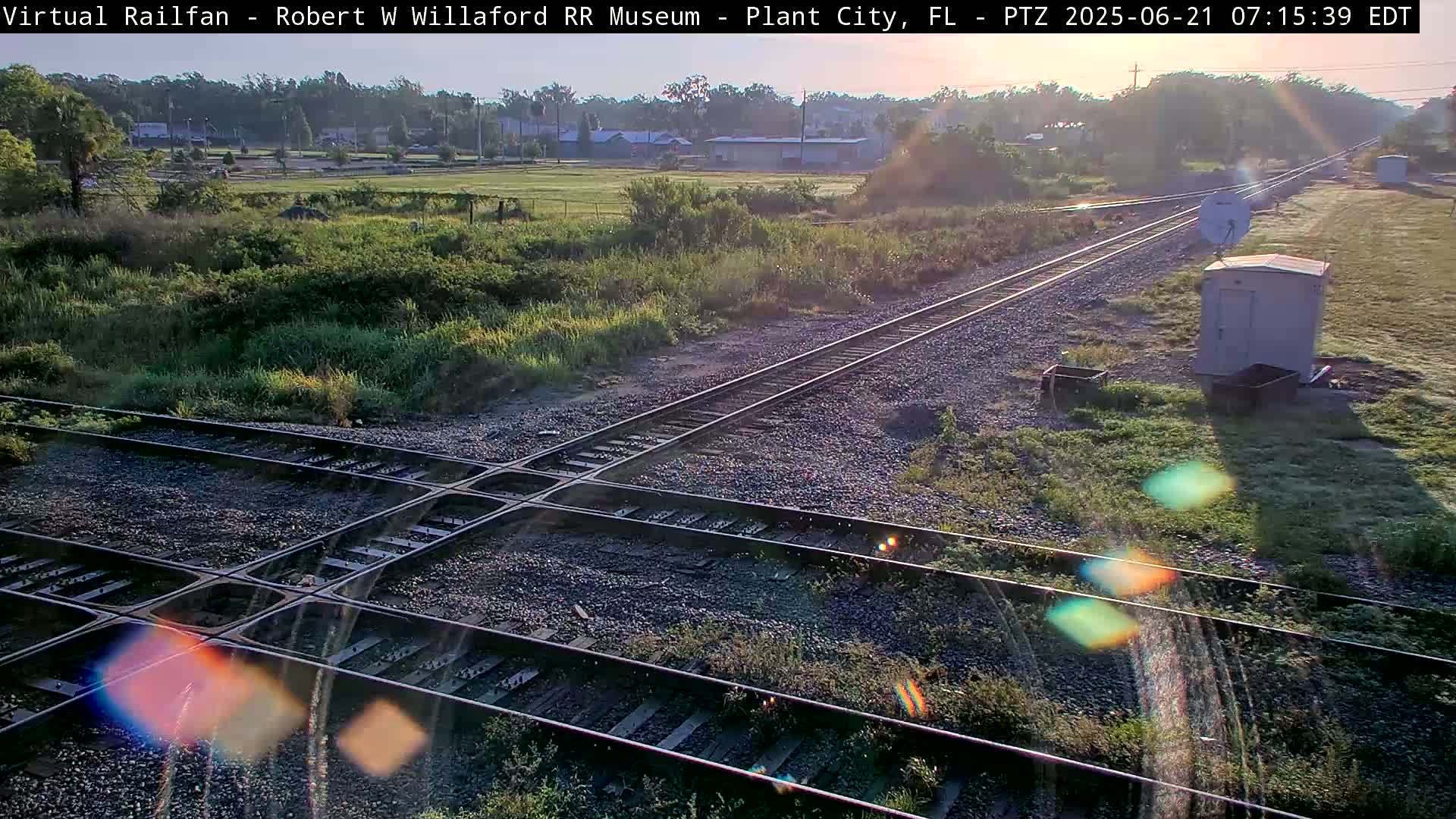 A sunlit view of a railroad crossing with vegetation and a small building nearby.