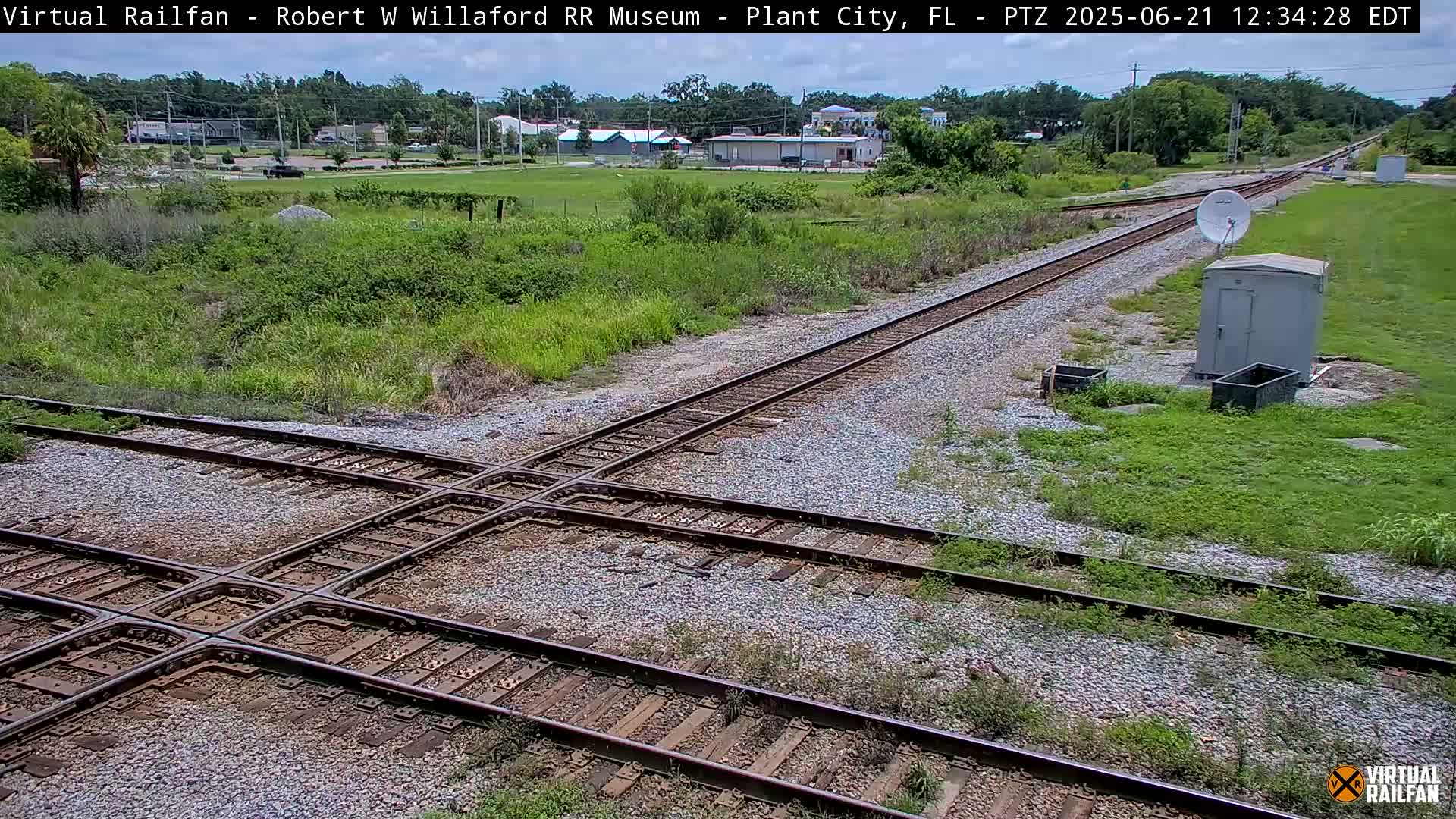 A railroad crossing with intersecting tracks is shown, surrounded by grassy areas and a small building, on a sunny day.