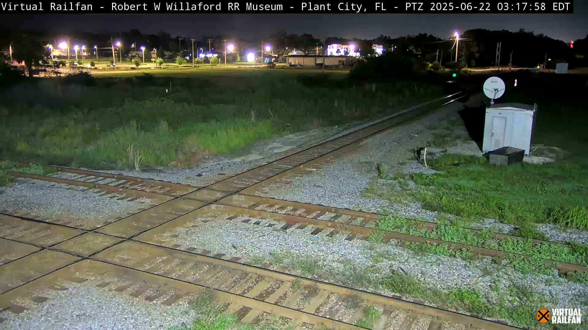 A nighttime view of a railroad crossing with intersecting tracks, surrounded by overgrown grass, and a small building with a satellite dish nearby.