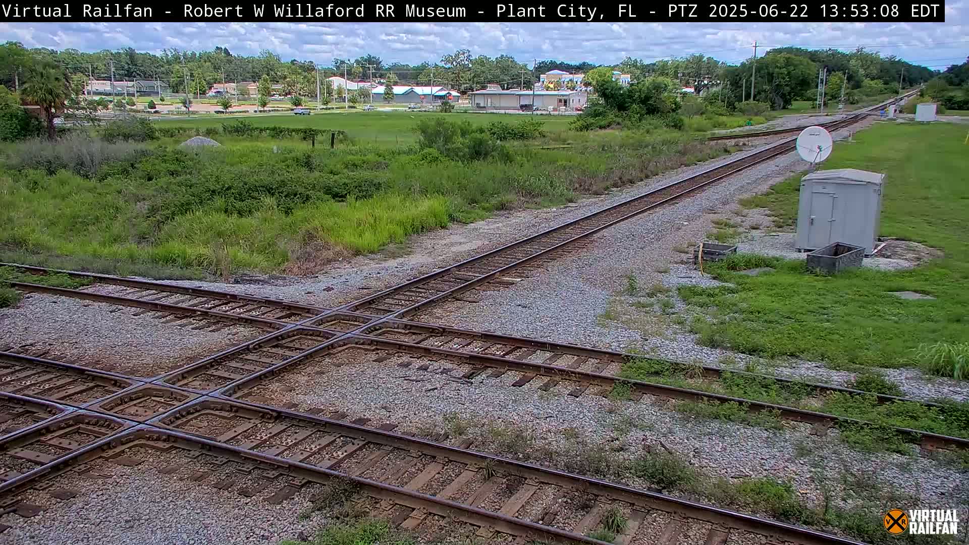 A railroad crossing with two sets of tracks intersecting, surrounded by grassy areas and a small building, under a partly cloudy sky.