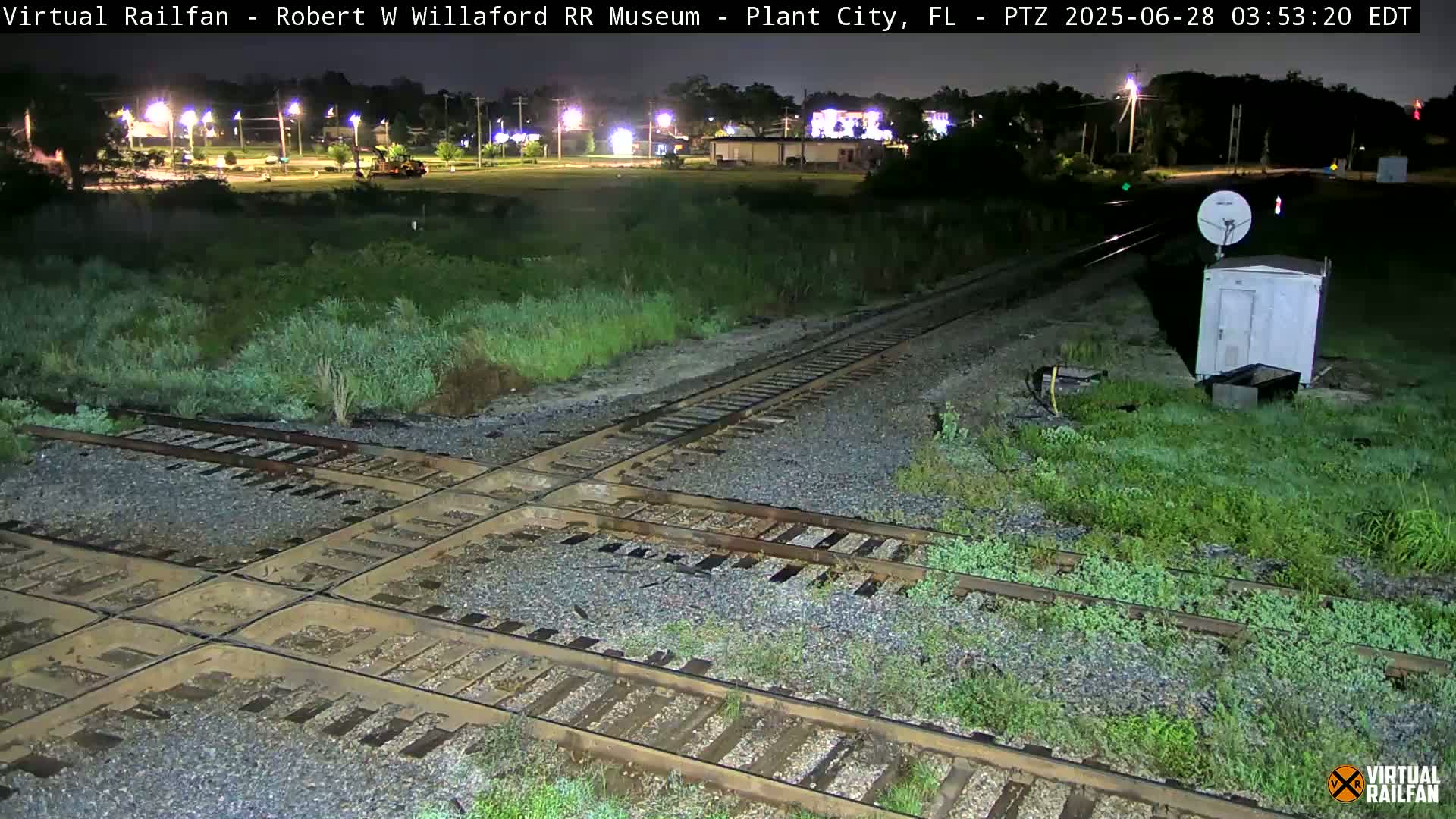 A nighttime view of a railroad crossing with overgrown vegetation, under dark, clear skies.