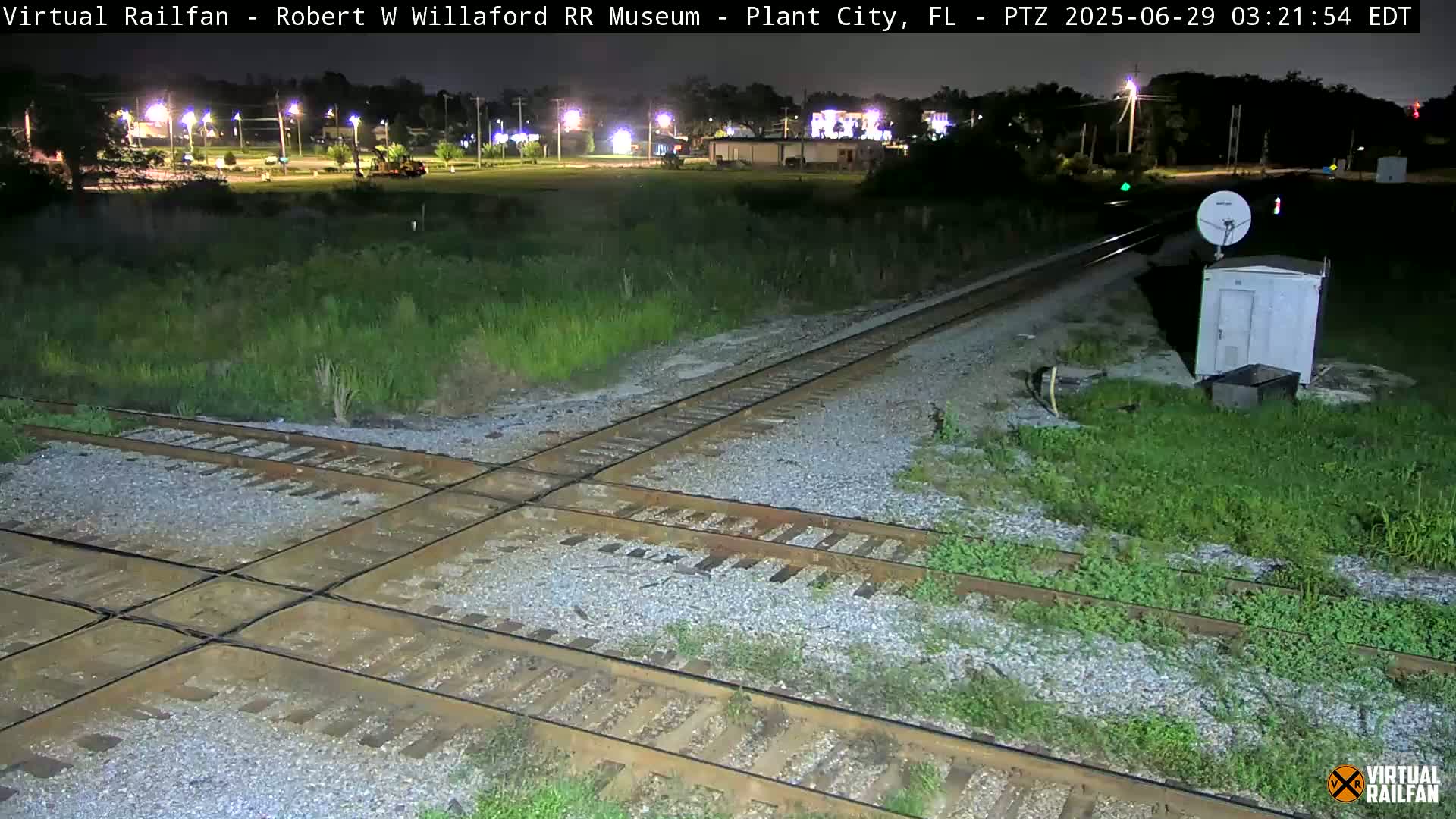 A nighttime view of a railroad crossing with a small building and satellite dish, surrounded by grassy areas and under a clear sky.