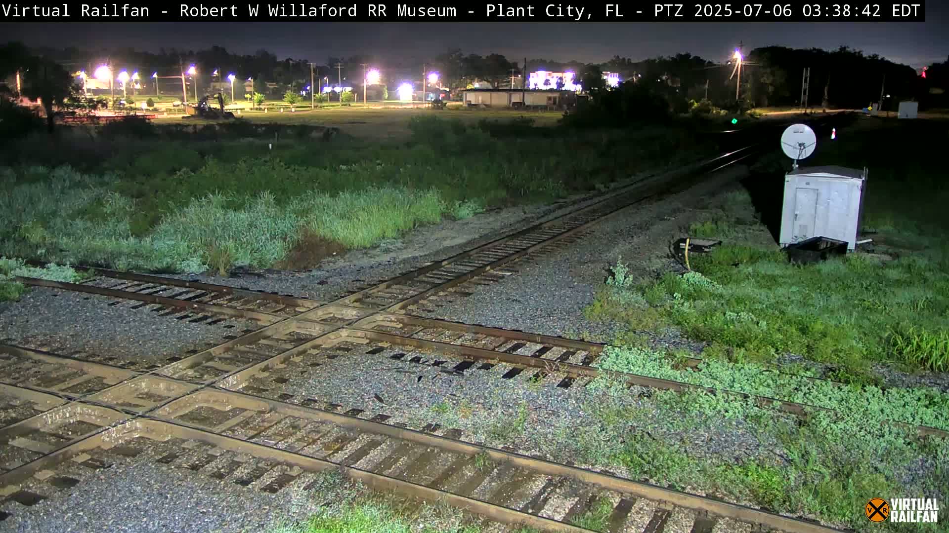 A nighttime view of a railroad crossing with several tracks intersecting, surrounded by overgrown vegetation and a small building, showing signs of recent rain.