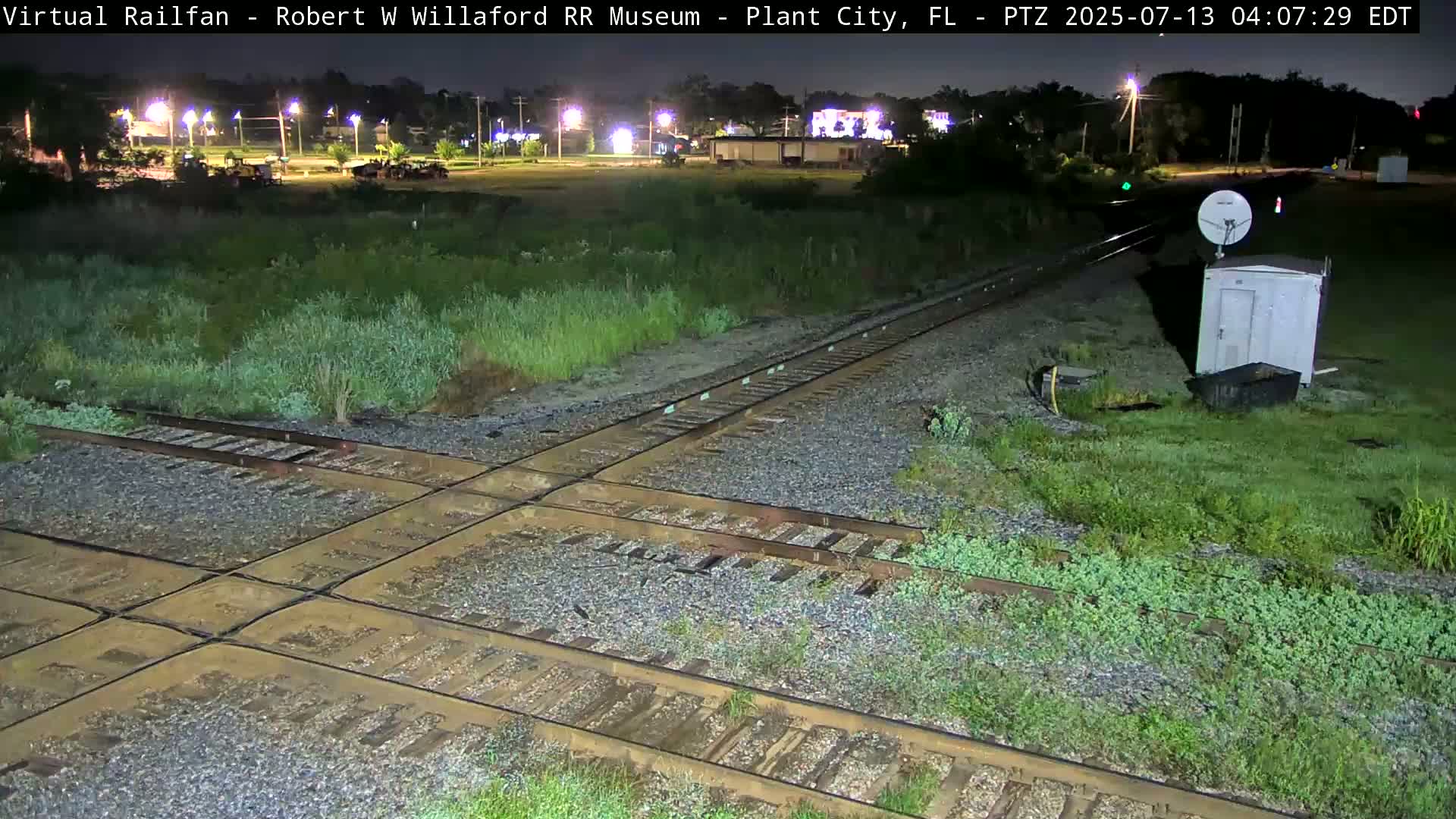 A nighttime view of a railroad crossing with overgrown vegetation and a small utility shed near a satellite dish.