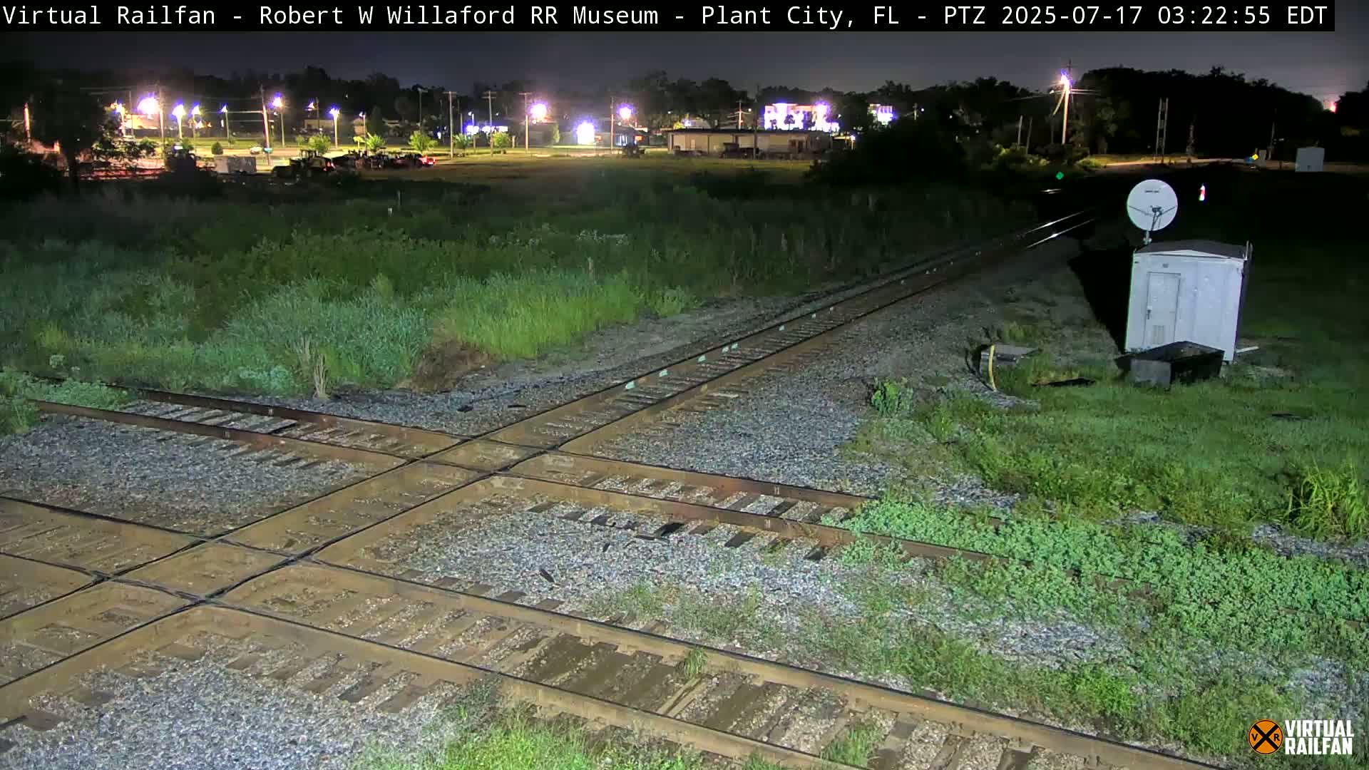 A nighttime view of a railroad crossing with intersecting tracks, surrounded by vegetation, under slightly wet conditions.