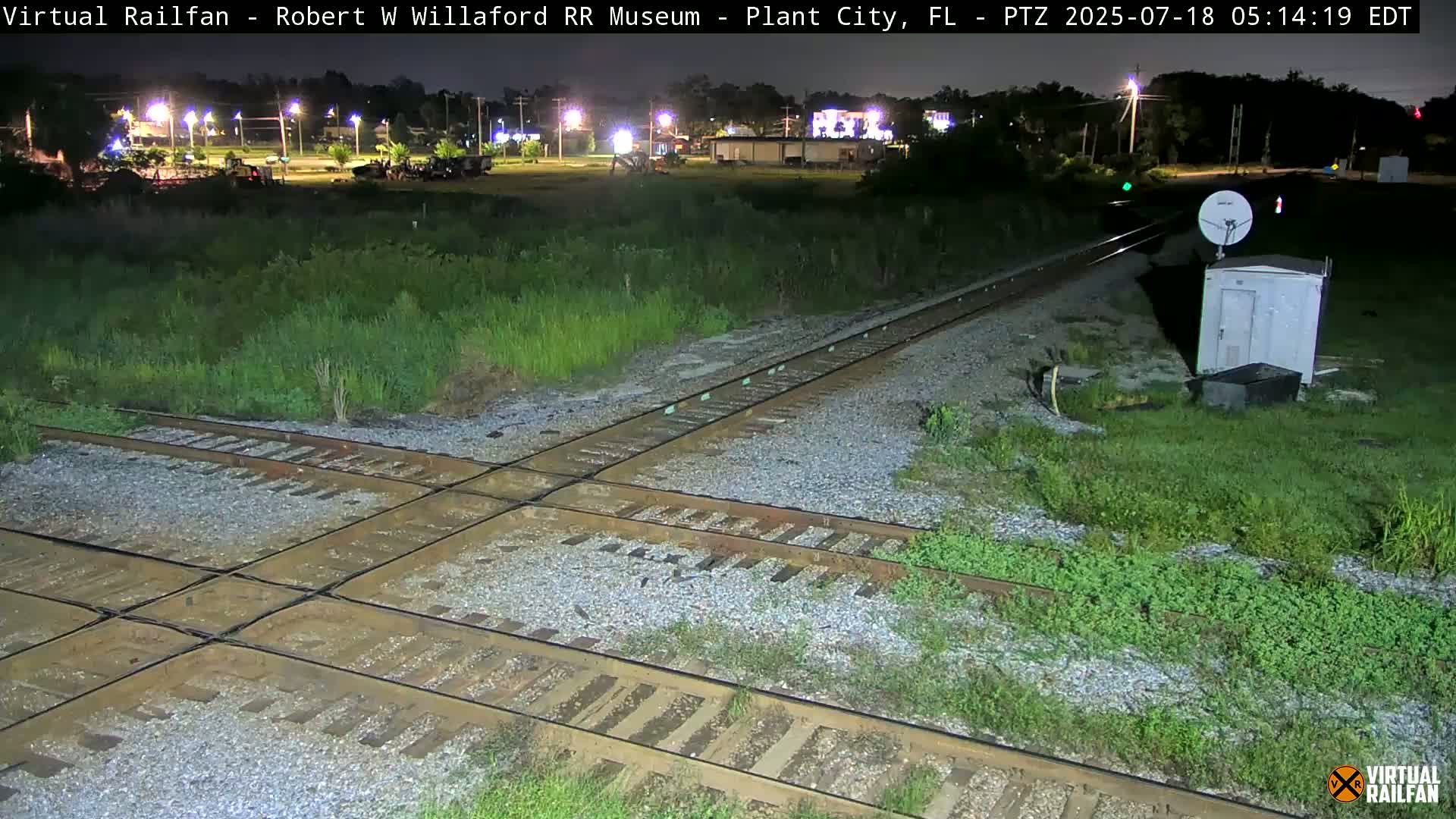 A nighttime view of a railroad crossing with intersecting tracks, surrounded by overgrown vegetation, under a clear sky.