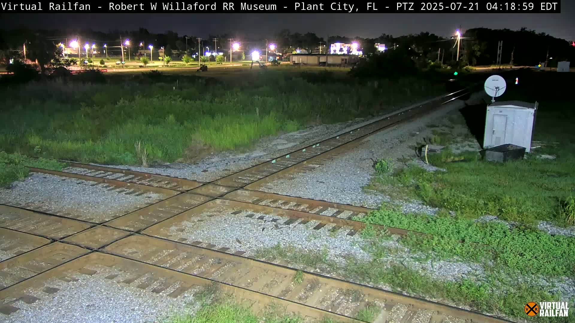 A nighttime view of a railroad crossing with train tracks intersecting, surrounded by overgrown vegetation under a clear sky.