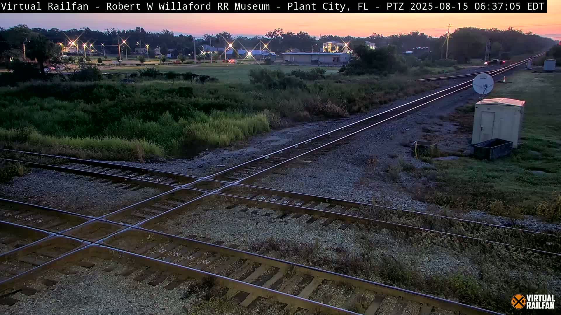 A nighttime view of a railroad crossing with train tracks intersecting, surrounded by overgrown vegetation under a clear sky.