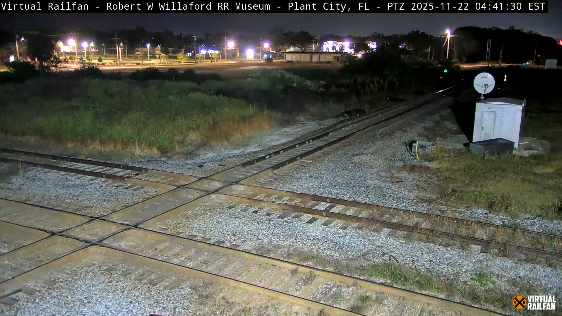 A clear nighttime view captures multiple railroad tracks intersecting over gravel and grass, with a utility box and satellite dish beside the tracks, and distant city lights illuminating the horizon.