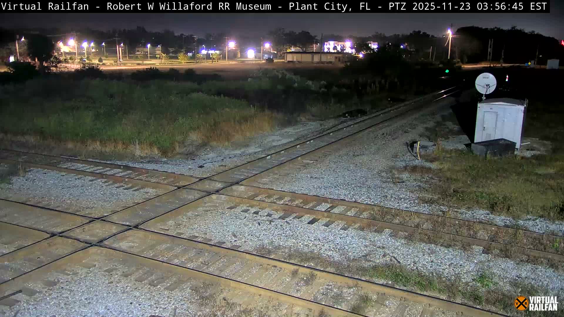 A complex intersection of multiple railroad tracks dominates the foreground, flanked by overgrown grass and a utility box with a satellite dish, under a clear night sky with distant city lights illuminating the horizon.
