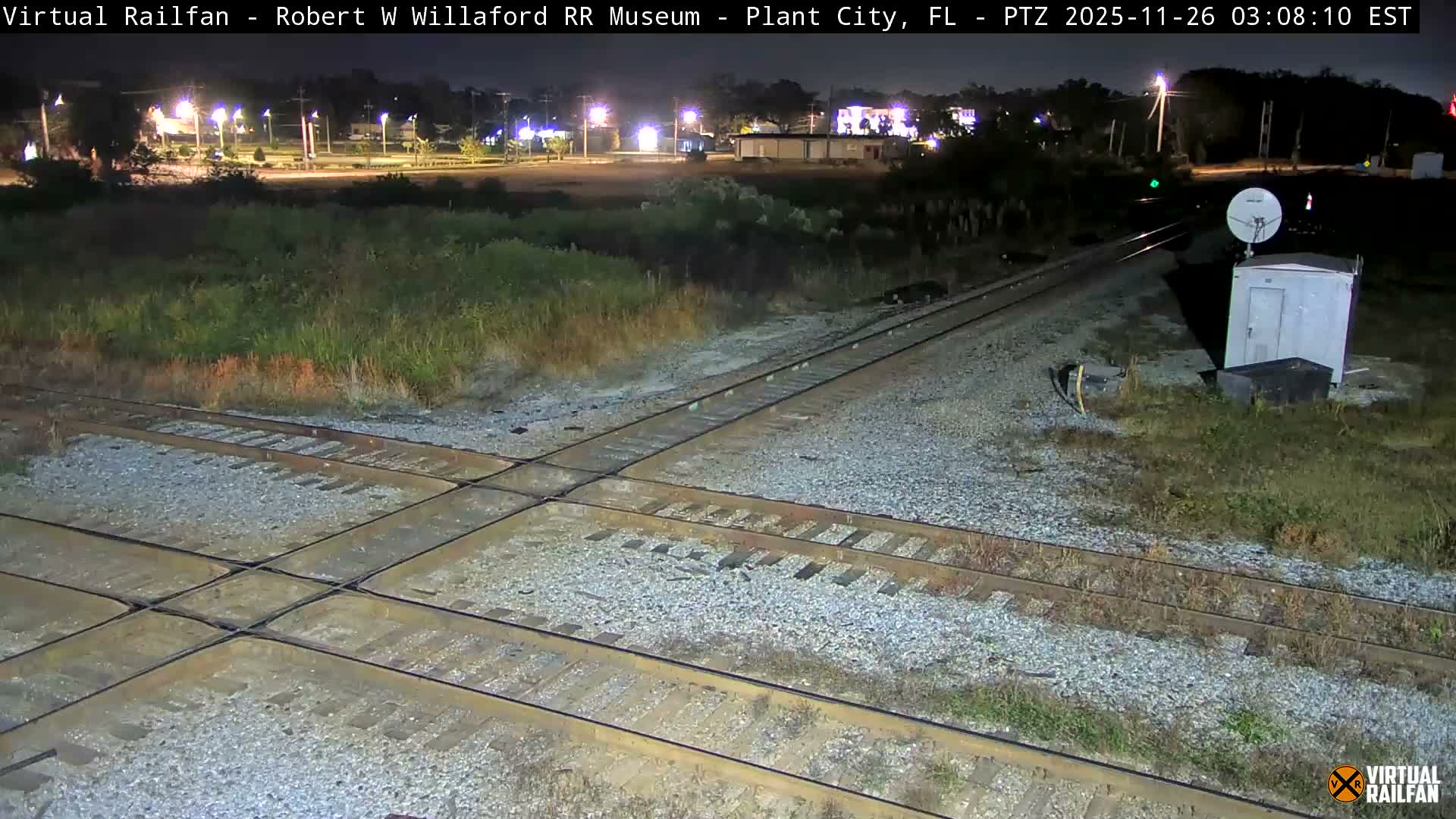 A clear nighttime image shows a railway crossing with multiple tracks surrounded by gravel and sparse vegetation, featuring a utility box with a satellite dish, a green signal light, and distant streetlights illuminating buildings in the background.