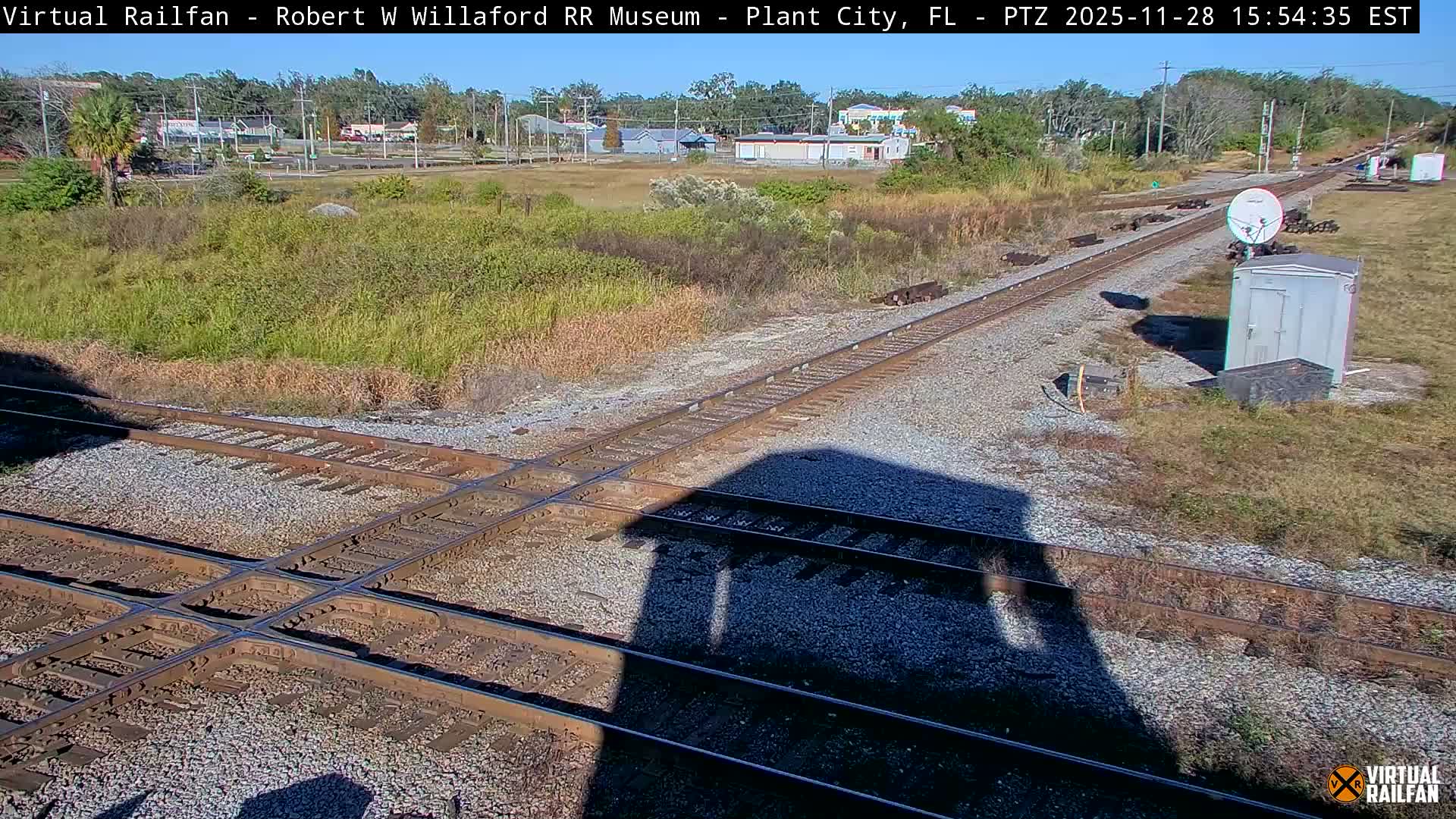 Under a clear, sunny sky, a prominent railway diamond crossing dominates the gravel-strewn foreground, with multiple tracks converging and diverging as they extend through grassy fields towards distant buildings and trees.