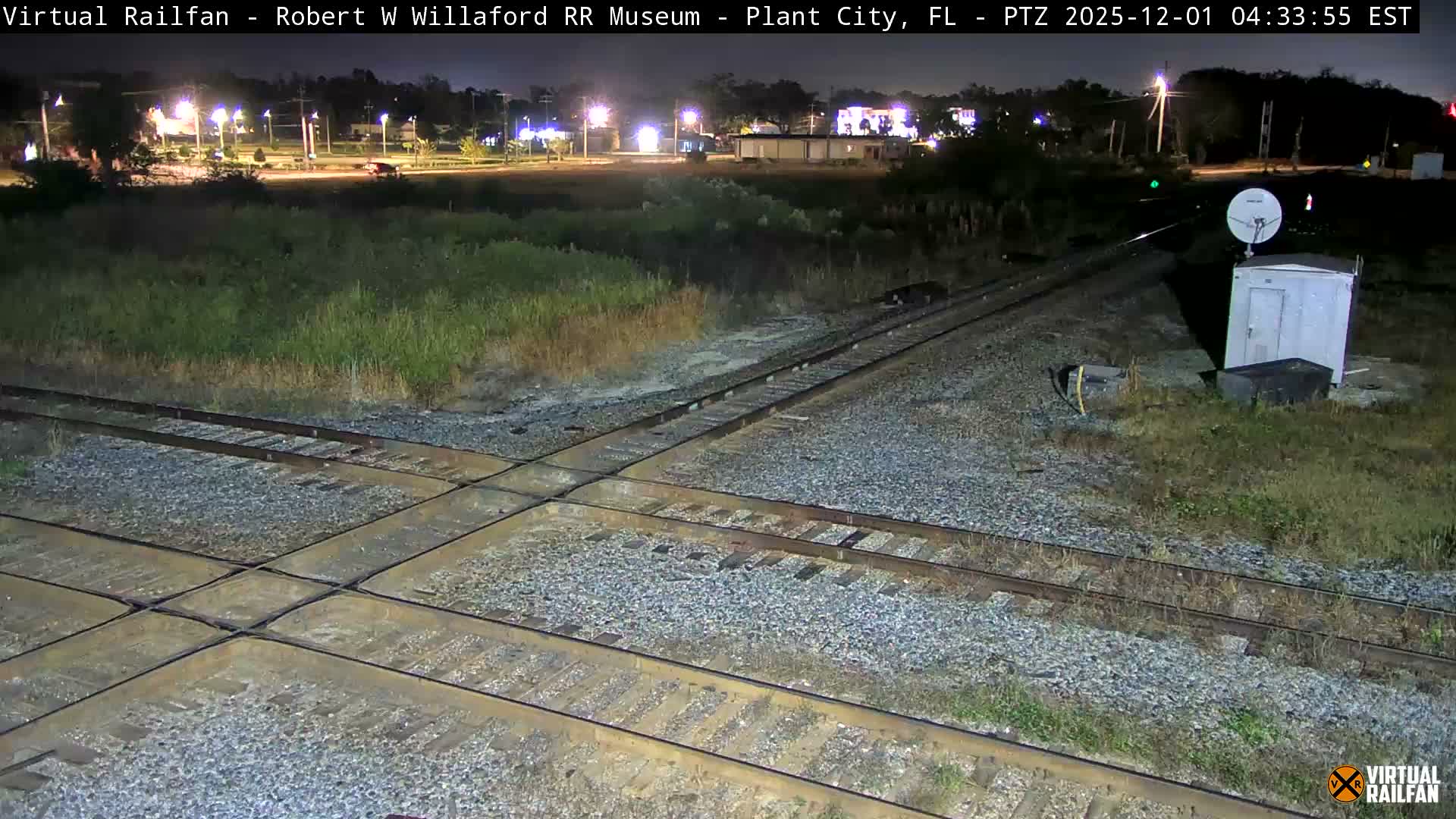 At night, multiple railroad tracks intersect amidst grassy terrain in the foreground, with a utility box and satellite dish to the right, and distant streetlights illuminating buildings under a clear sky.