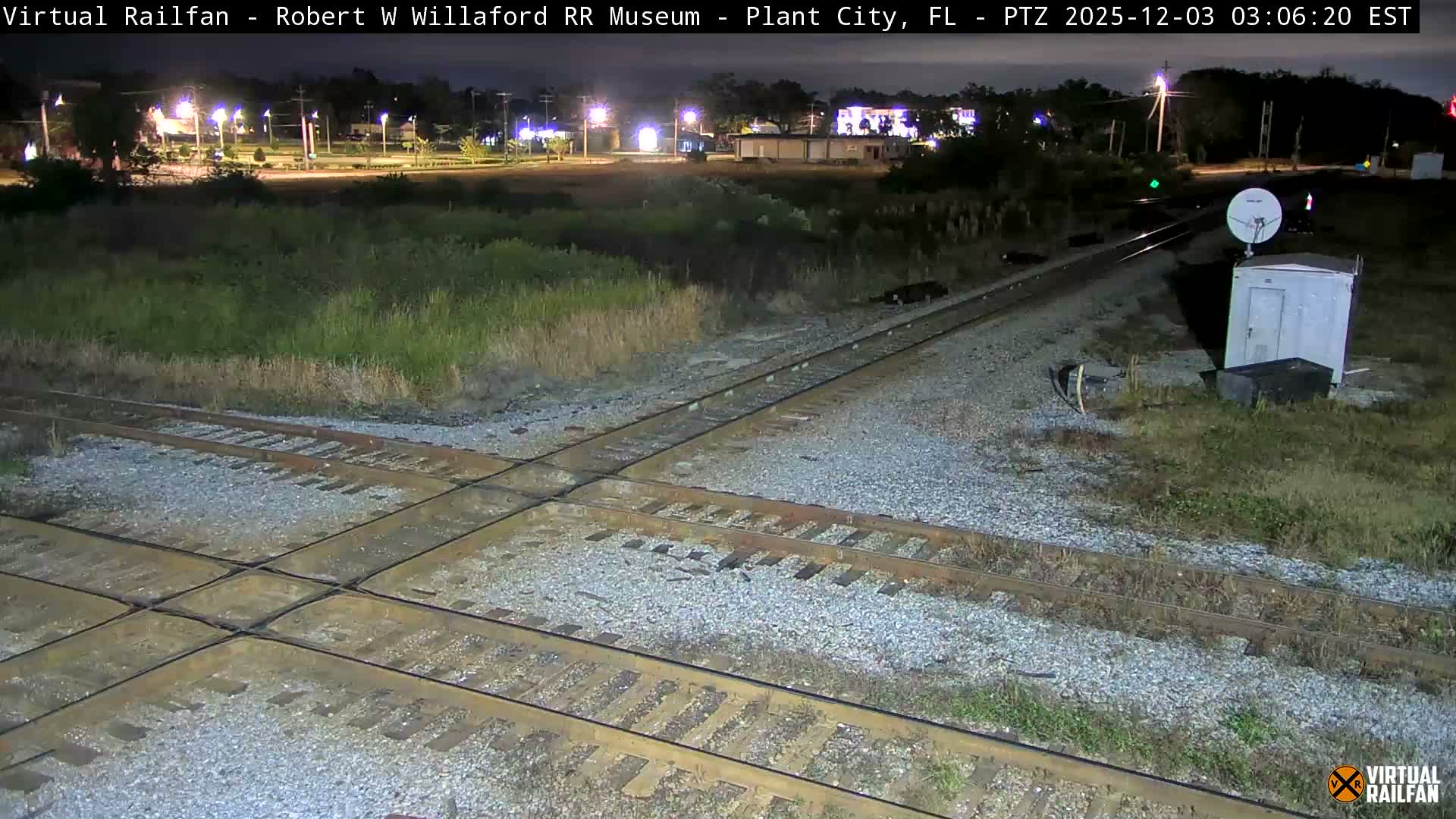 At night, a clear outdoor scene shows railroad tracks intersecting in the foreground, bordered by a utility shed with a satellite dish to the right, and distant urban lights illuminating the dark sky.