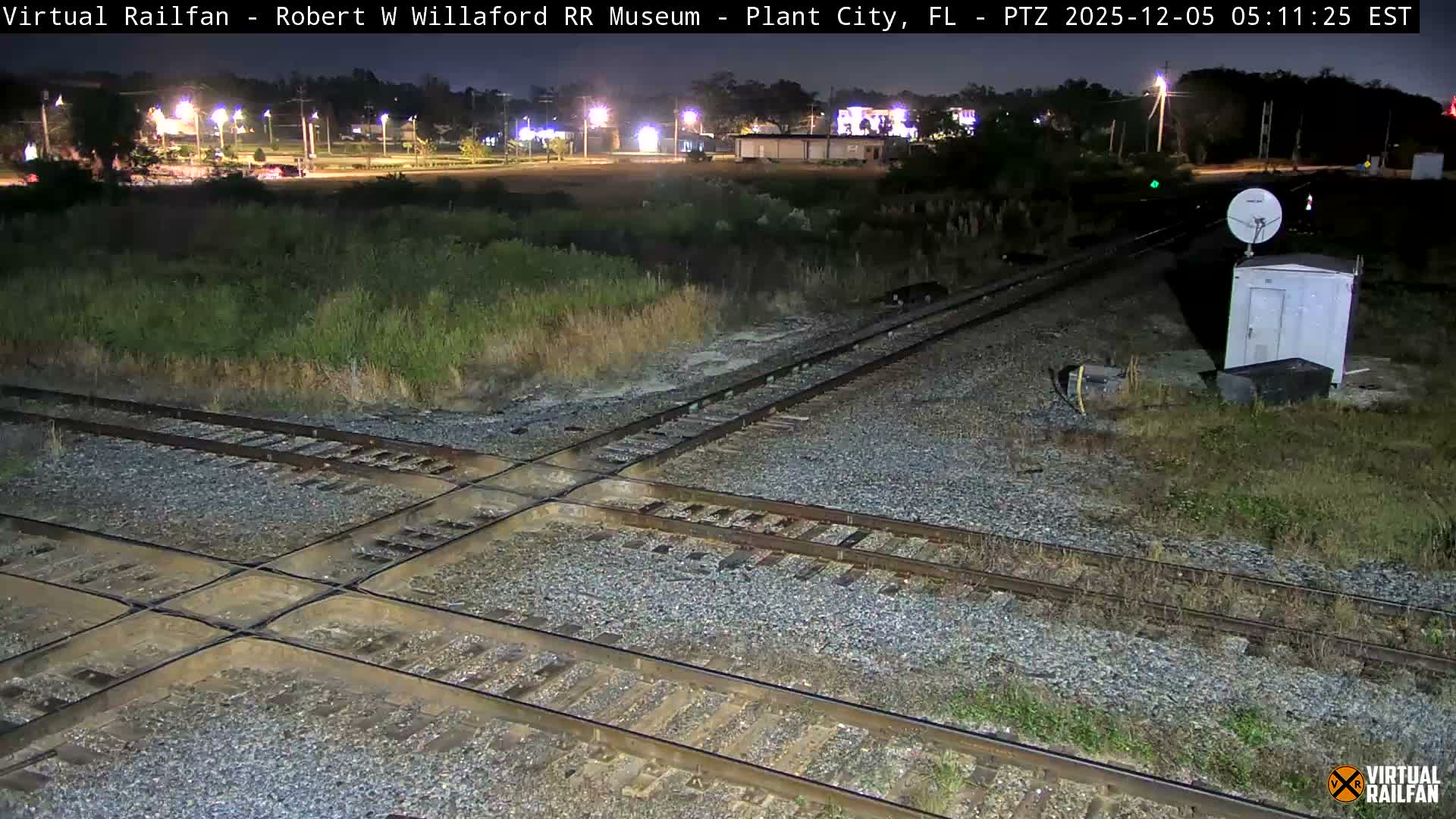 A clear, dark night illuminates a complex railroad track intersection, with a white utility shed and satellite dish positioned on the right, and various distant lights scattered across the background.