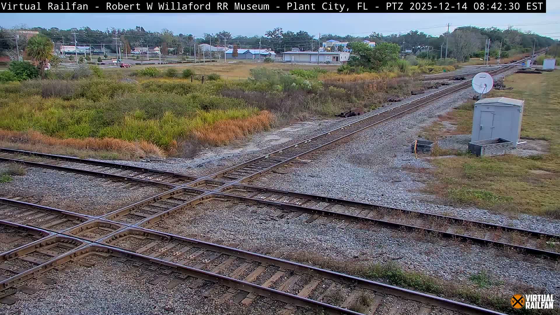A clear, dark night illuminates a complex railroad track intersection, with a white utility shed and satellite dish positioned on the right, and various distant lights scattered across the background.