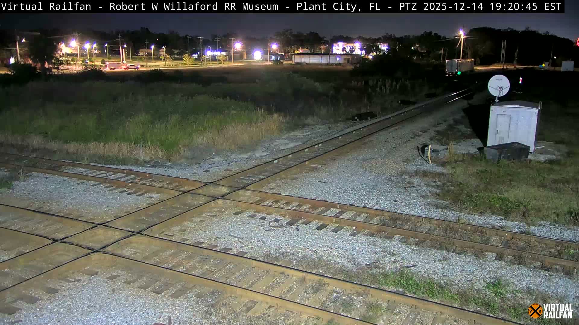 A clear, dark night illuminates a complex railroad track intersection, with a white utility shed and satellite dish positioned on the right, and various distant lights scattered across the background.