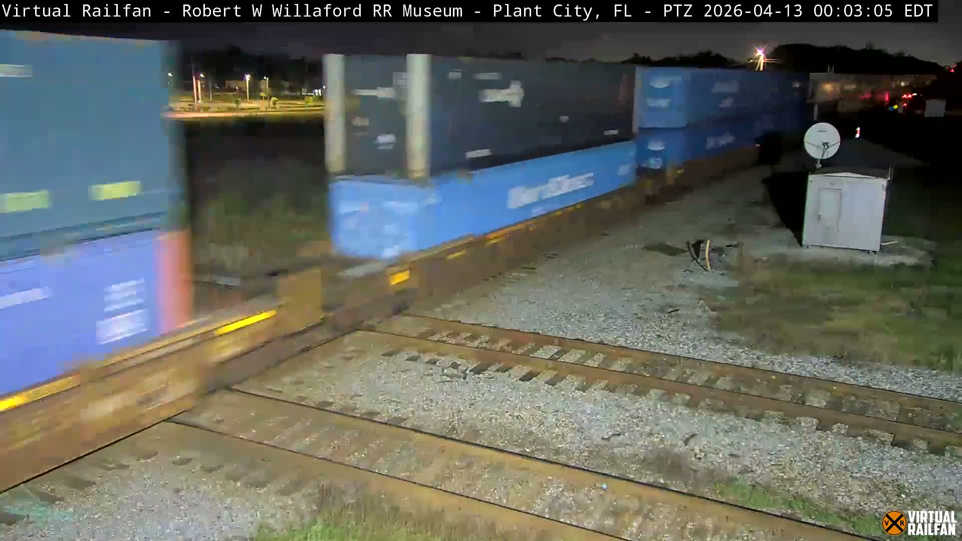 A clear, dark night illuminates a complex railroad track intersection, with a white utility shed and satellite dish positioned on the right, and various distant lights scattered across the background.