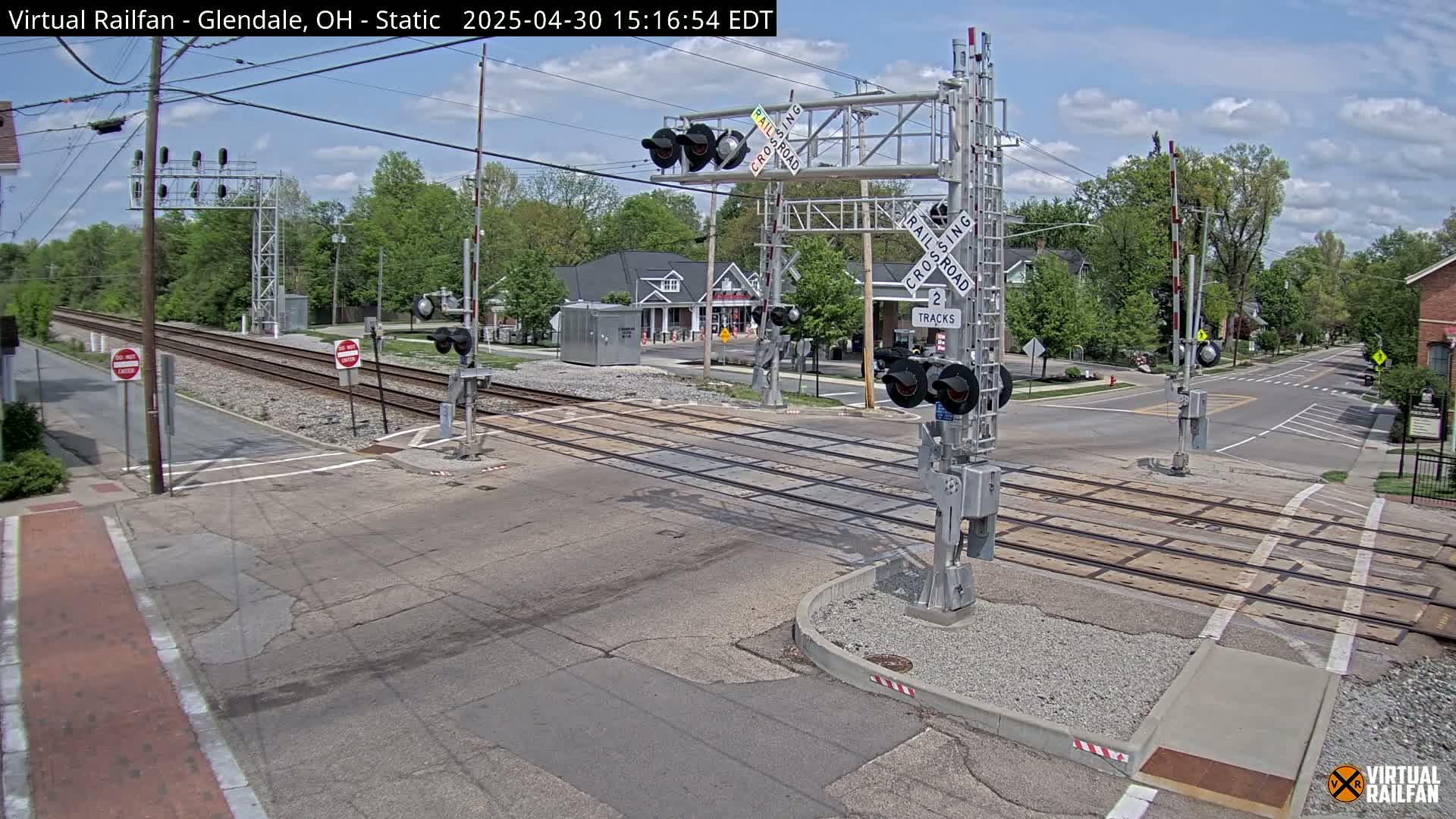 A sunny day shows a railroad crossing with lowered crossing gates and signals, intersecting a road with buildings and trees visible in the background.