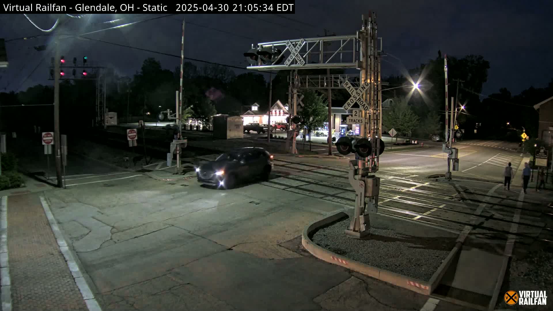 At night, a dark-colored SUV drives through a lightly trafficked railroad crossing under illuminated crossing gates.