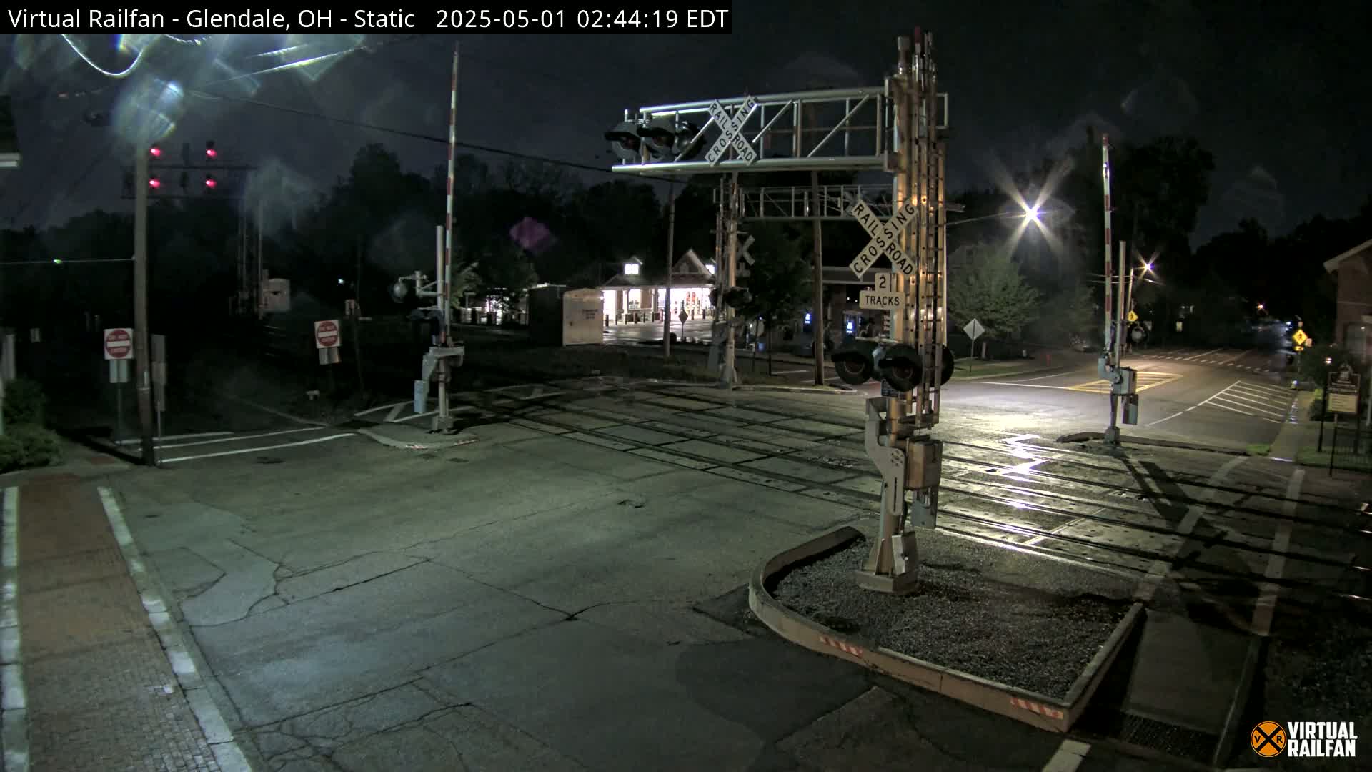 A nighttime view of a railroad crossing with the gates down, illuminated by streetlights and the glow of a nearby building.