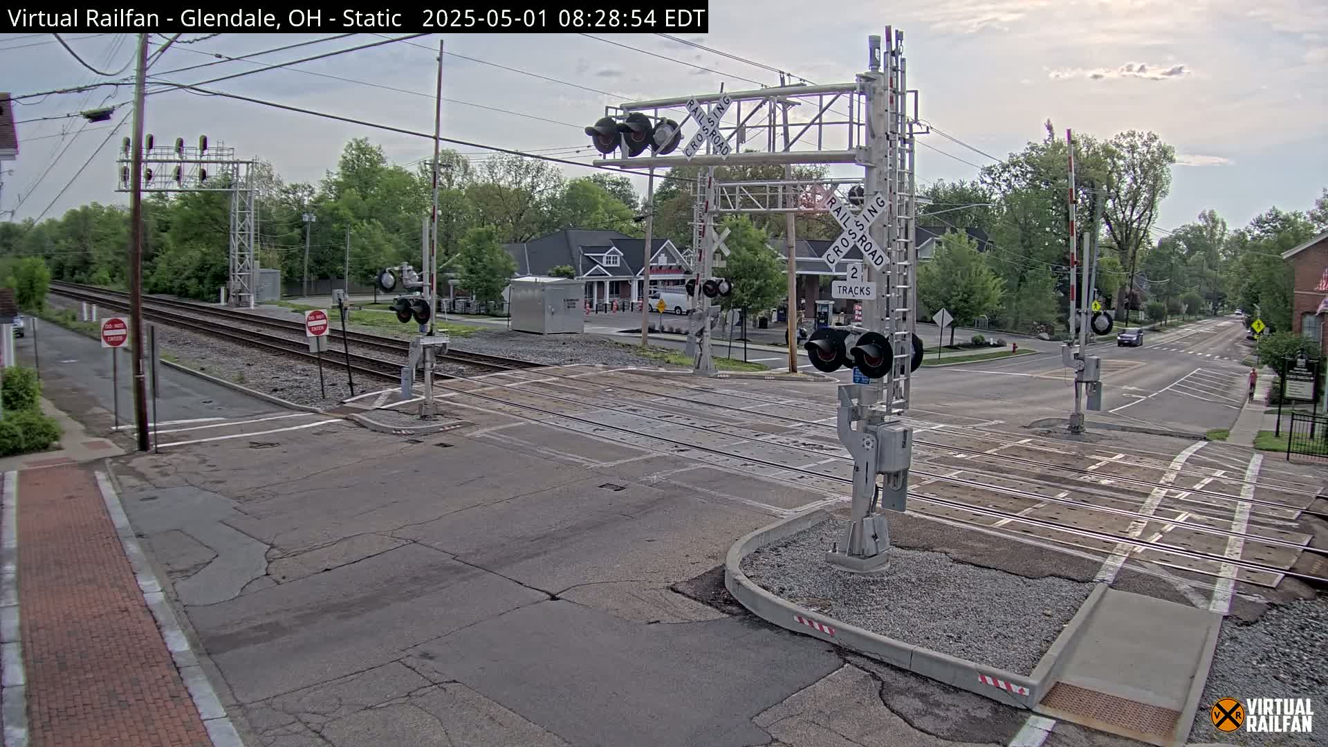 A railroad crossing with lowered gates and flashing lights is situated at a street intersection on an overcast day, with trees and buildings visible in the background.