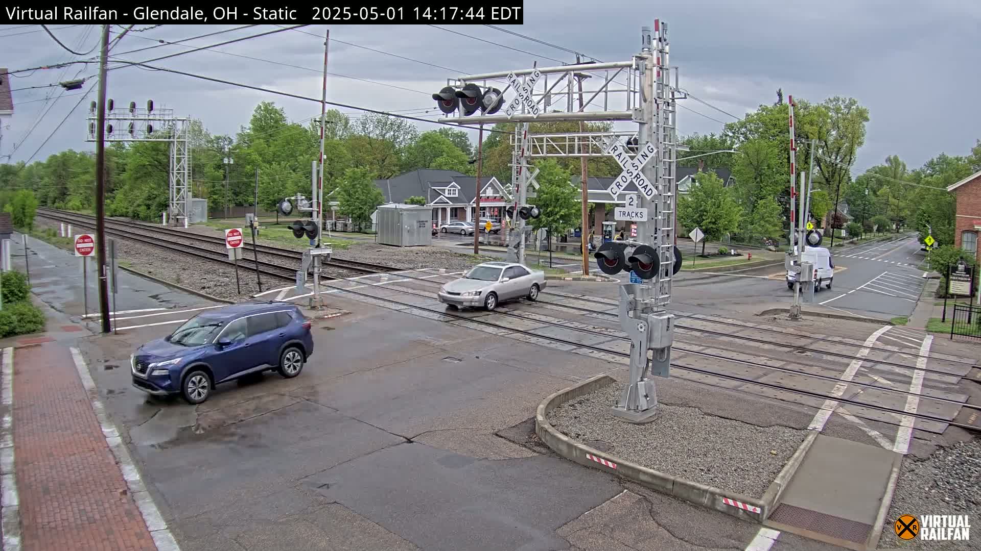 A wet street intersects with a railroad crossing, where several vehicles are stopped or slowly passing through, under an overcast sky.