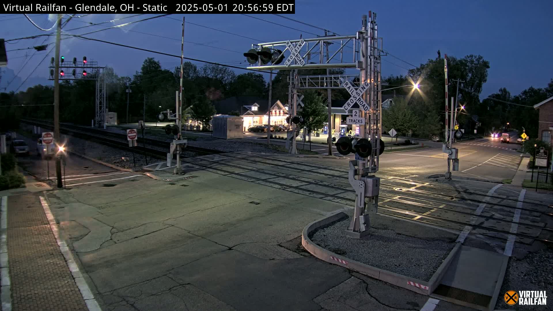 A nighttime view of a railroad crossing with lowered gates, traffic signals, and a nearly empty street under a twilight sky.