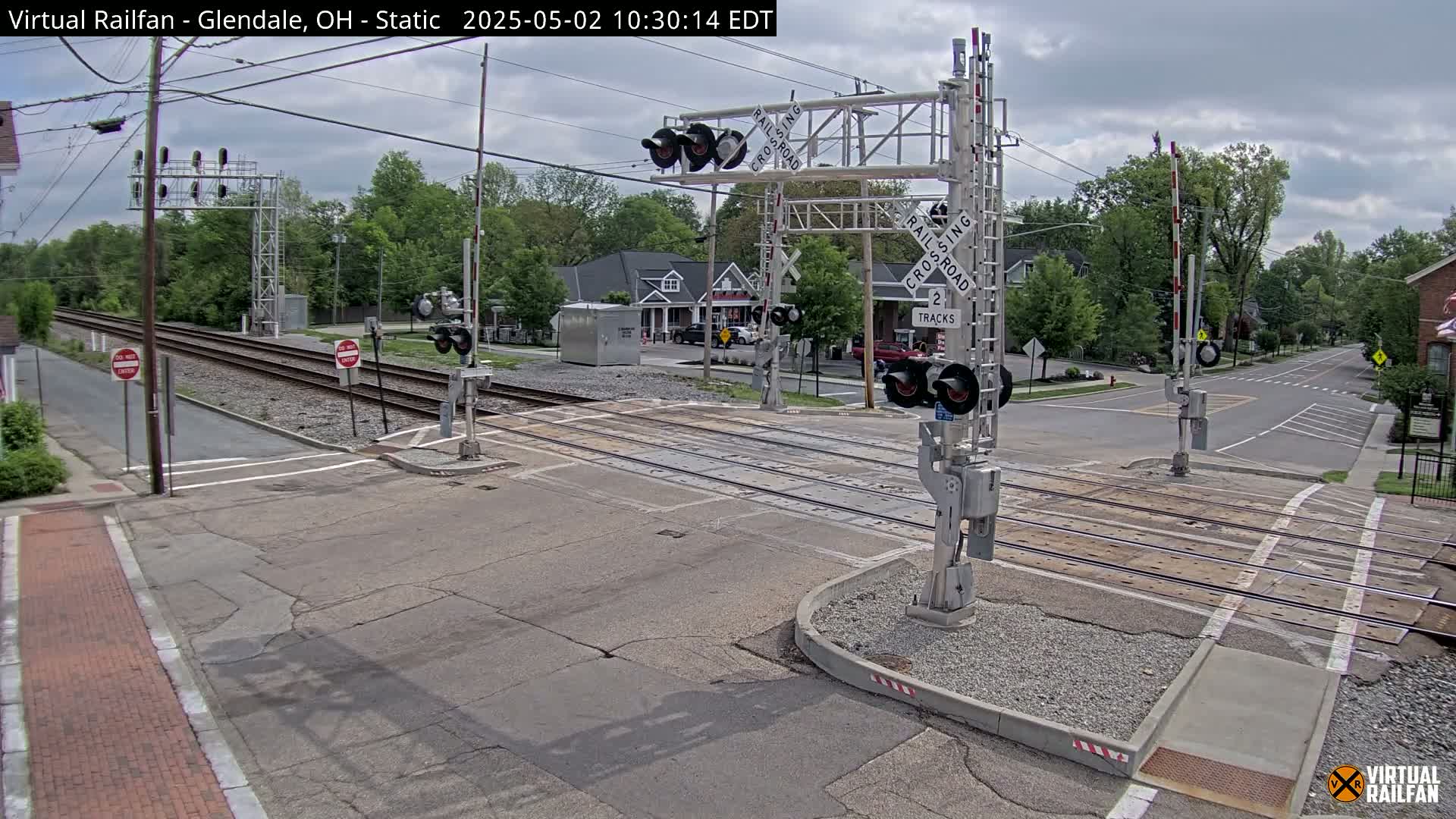 A railroad crossing with lowered gates is shown on an overcast day, with trees and buildings visible in the background.