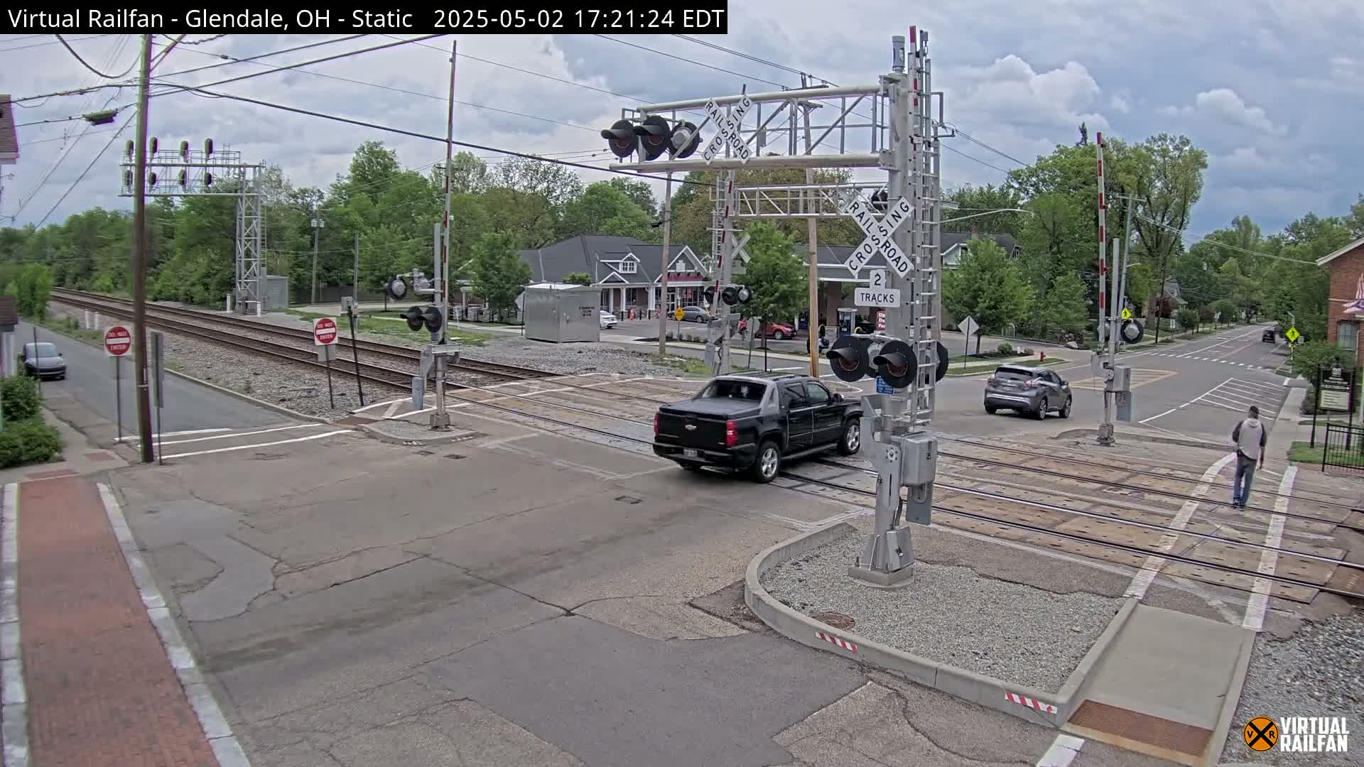 A partly cloudy day shows a railroad crossing with a dark-colored pickup truck and a silver SUV stopped at the crossing, a pedestrian walking by, and some buildings visible in the background.
