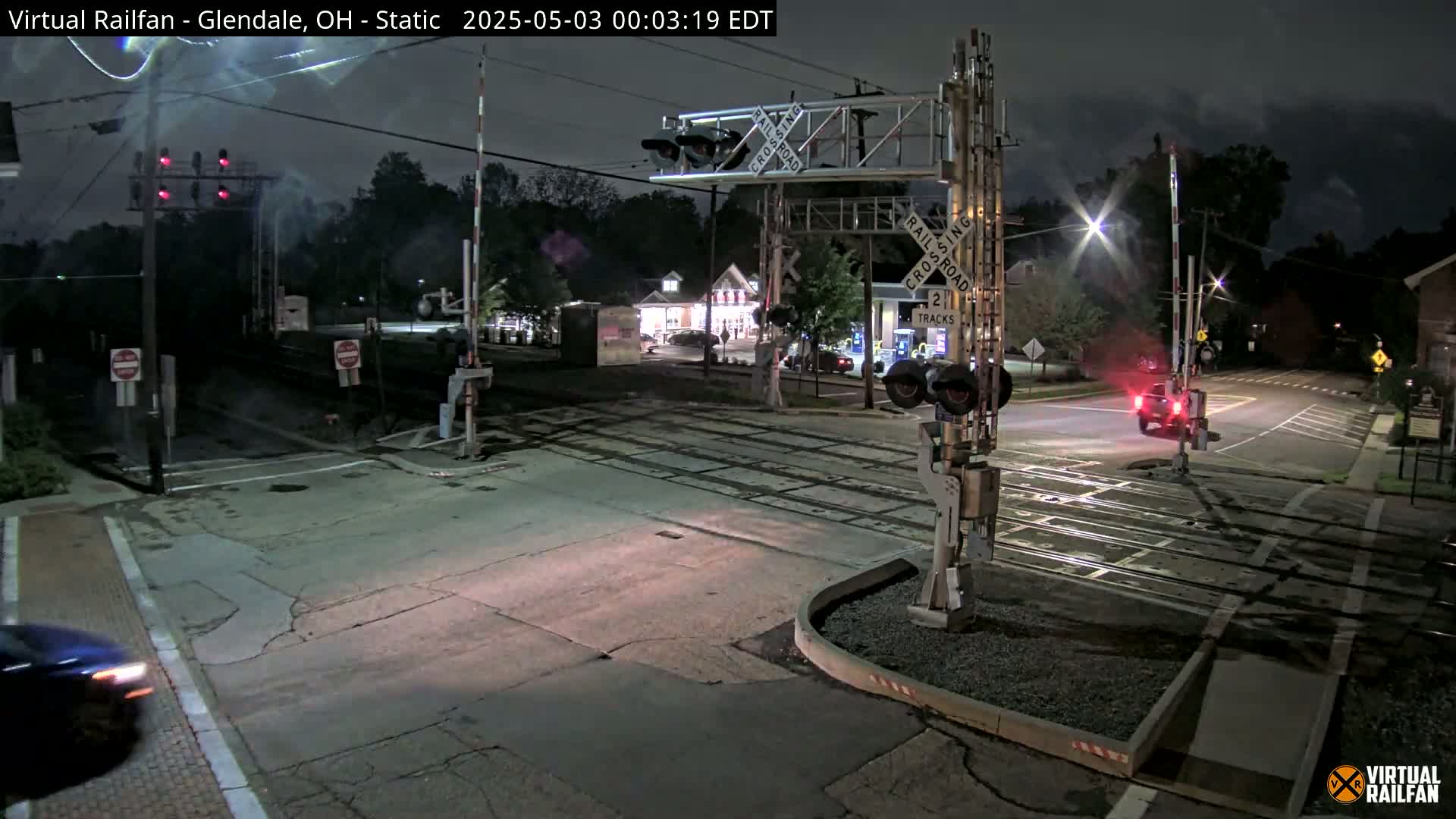 A nighttime view of a railroad crossing with a vehicle passing through, under illuminated streetlights and partly cloudy skies.