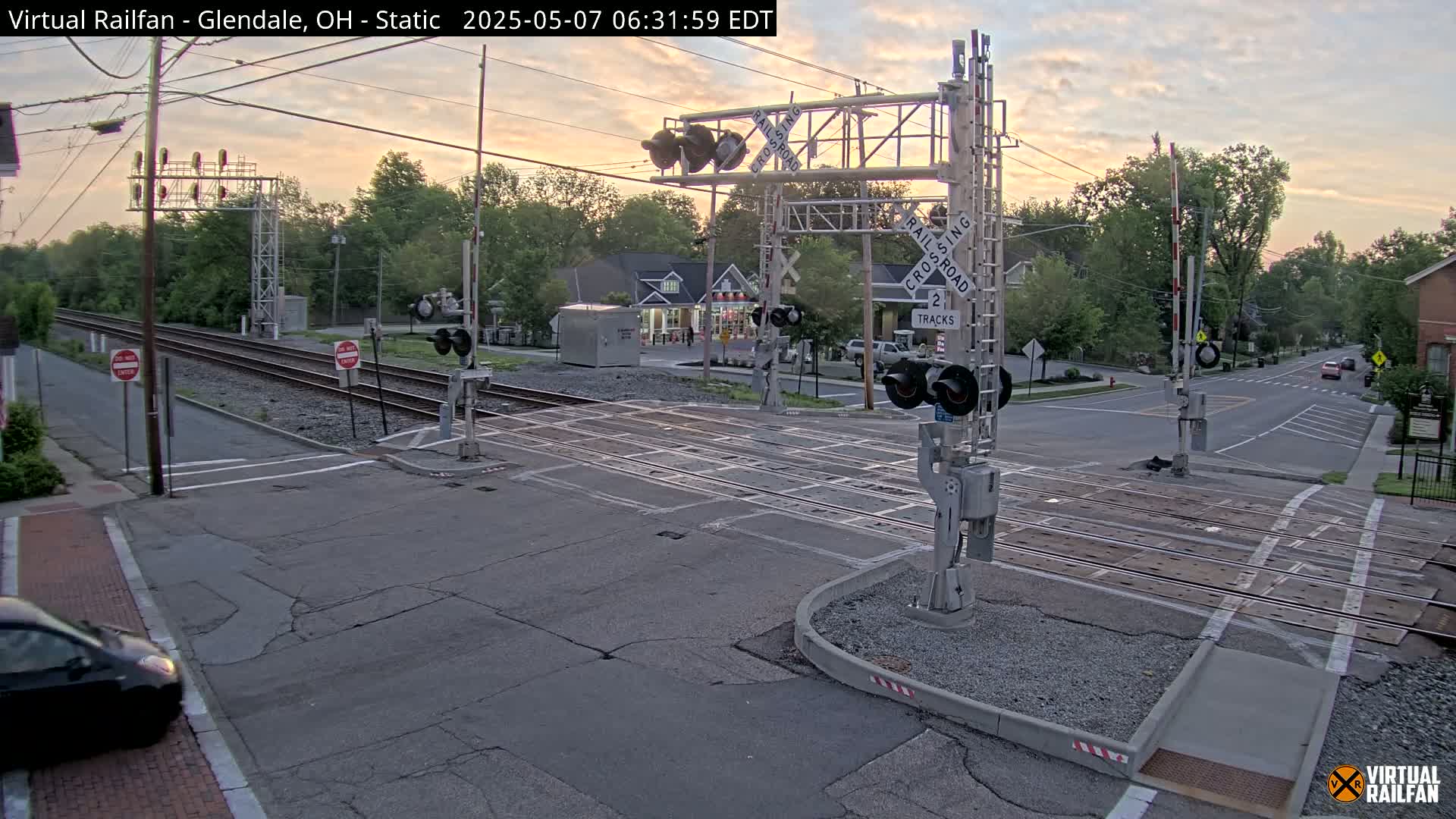 A railroad crossing with lowered gates, during early morning twilight, shows two sets of tracks intersecting a road in a residential area with trees and a small building visible in the background.