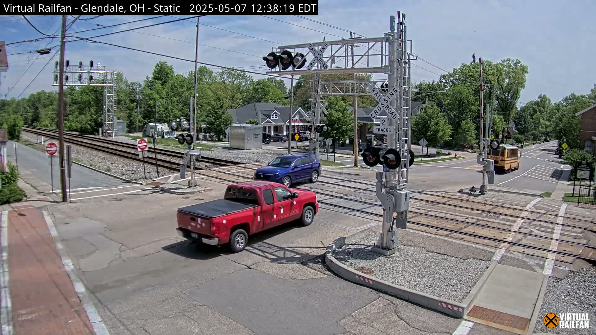 A red pickup truck and a blue SUV are crossing a railroad intersection under a sunny sky, with a school bus visible in the distance.