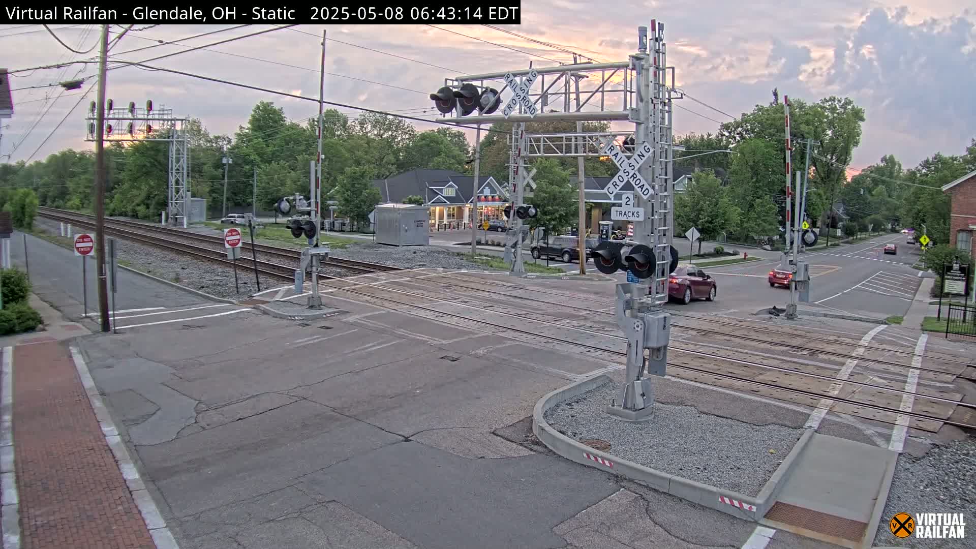 A quiet, overcast evening scene at a railroad crossing shows two sets of tracks intersecting a road, with a few cars visible and trees lining the streets.